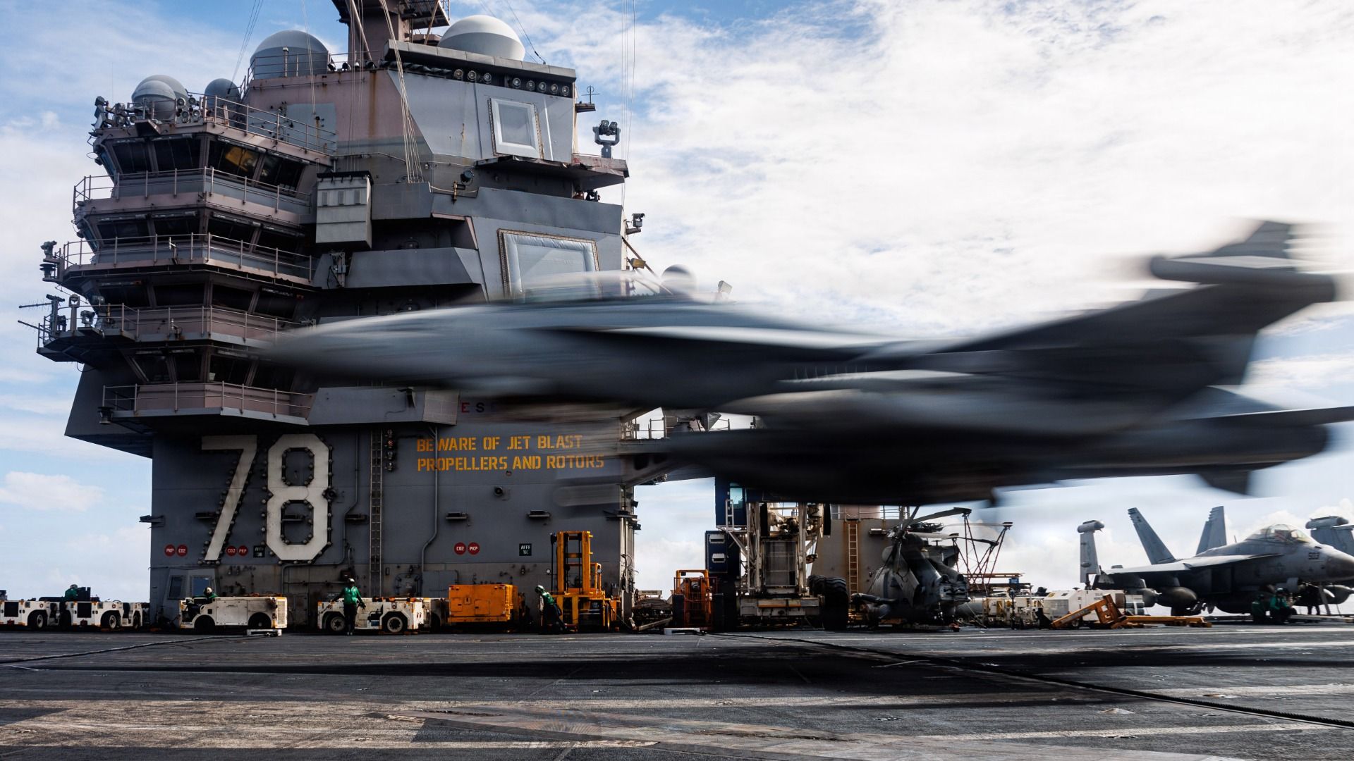 Blurred jet aircraft landing on an aircraft carrier with the number 78 visible on the tower. Workers and equipment are on the deck under a cloudy sky.