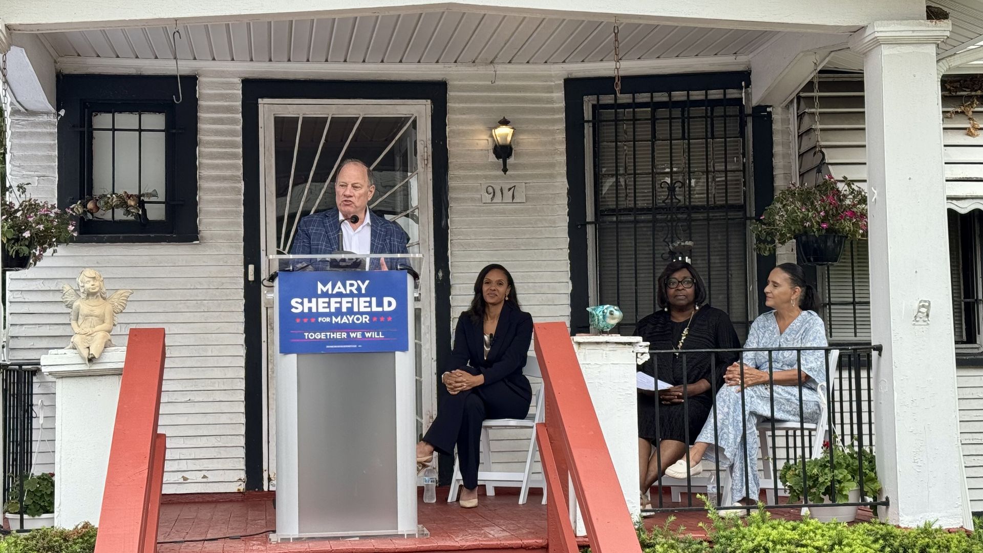 Mike Duggan speaks at a lectern with a "Mary Sheffield for Mayor" sign on it. Mary Sheffield sits nearby