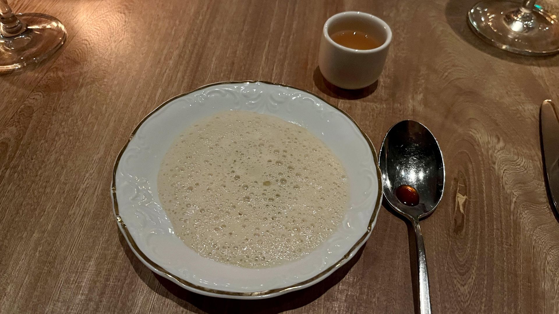 White bowl with a foamy beige soup on a wooden table, next to a spoon with a drop of red sauce and a small white cup with amber liquid.