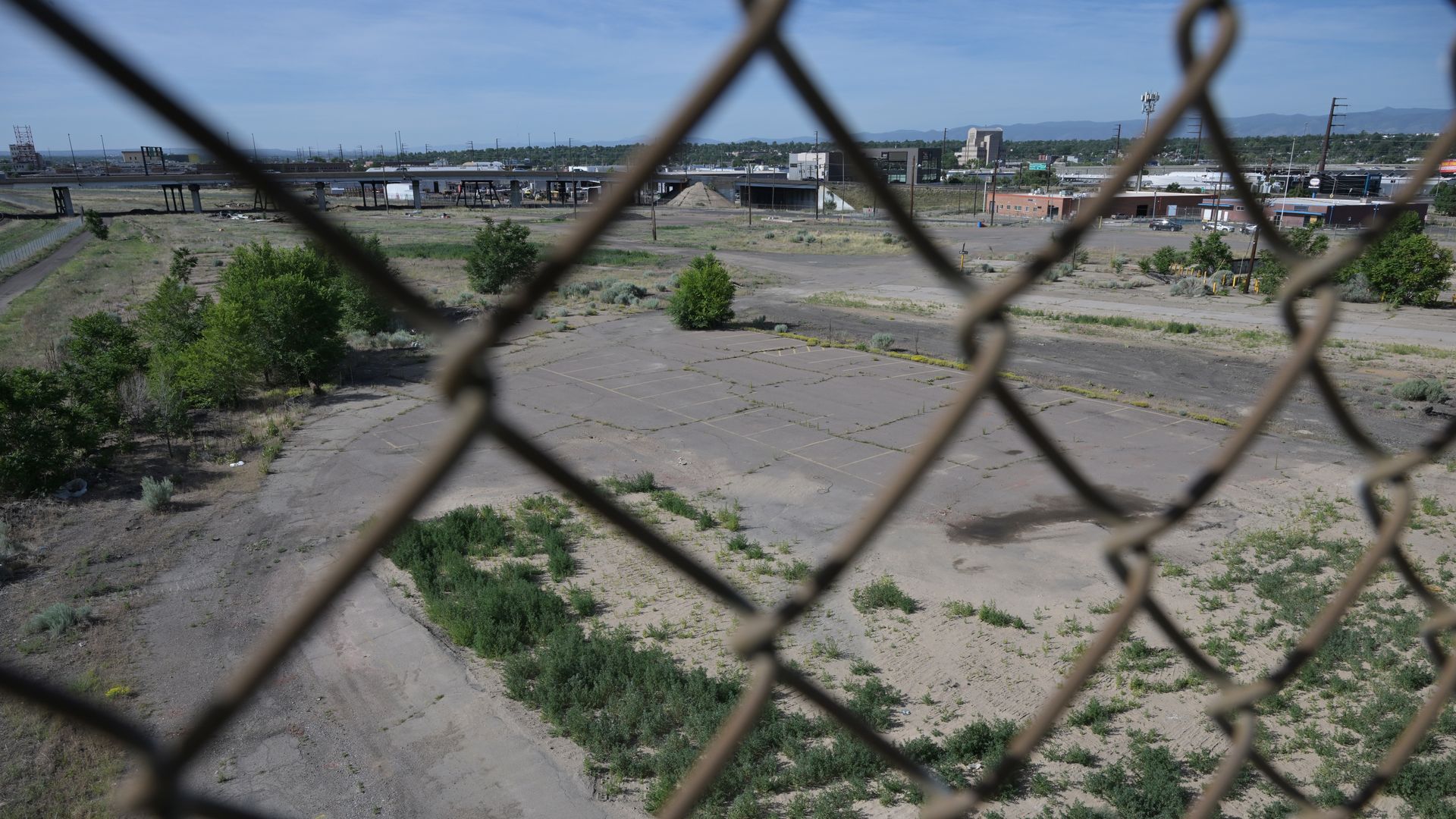 View of an abandoned industrial lot seen through a blurred chain-link fence, cracked pavement, sparse vegetation, distant highway overpasses, and low-rise buildings under a blue sky.