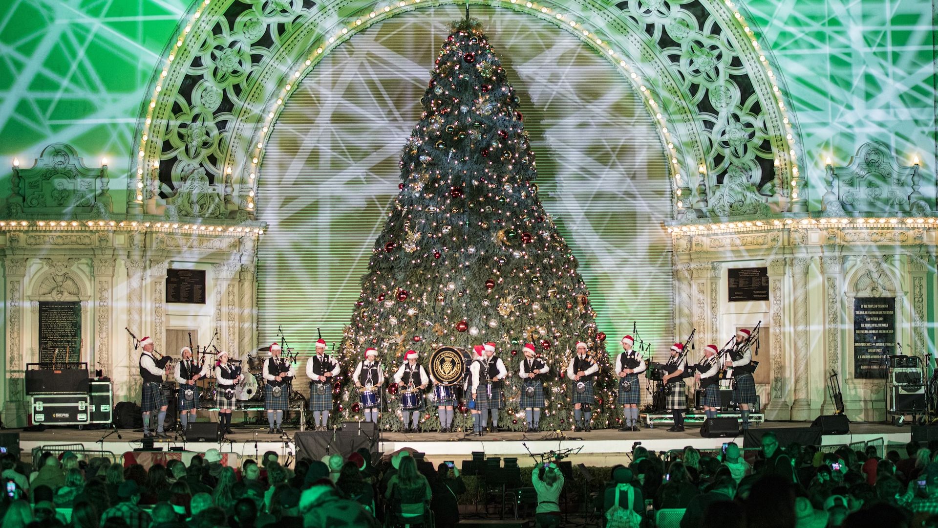 A Scottish pipe band performs on an outdoor stage in front of a large Christmas tree for a crowd.