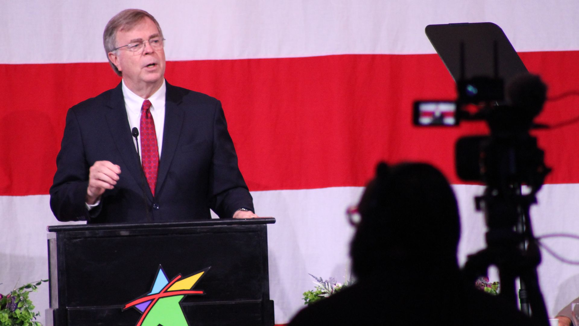 Man in a dark suit with red tie speaking at a podium with a colorful star logo, against a backdrop of red and white stripes, with camera and person silhouette in foreground.