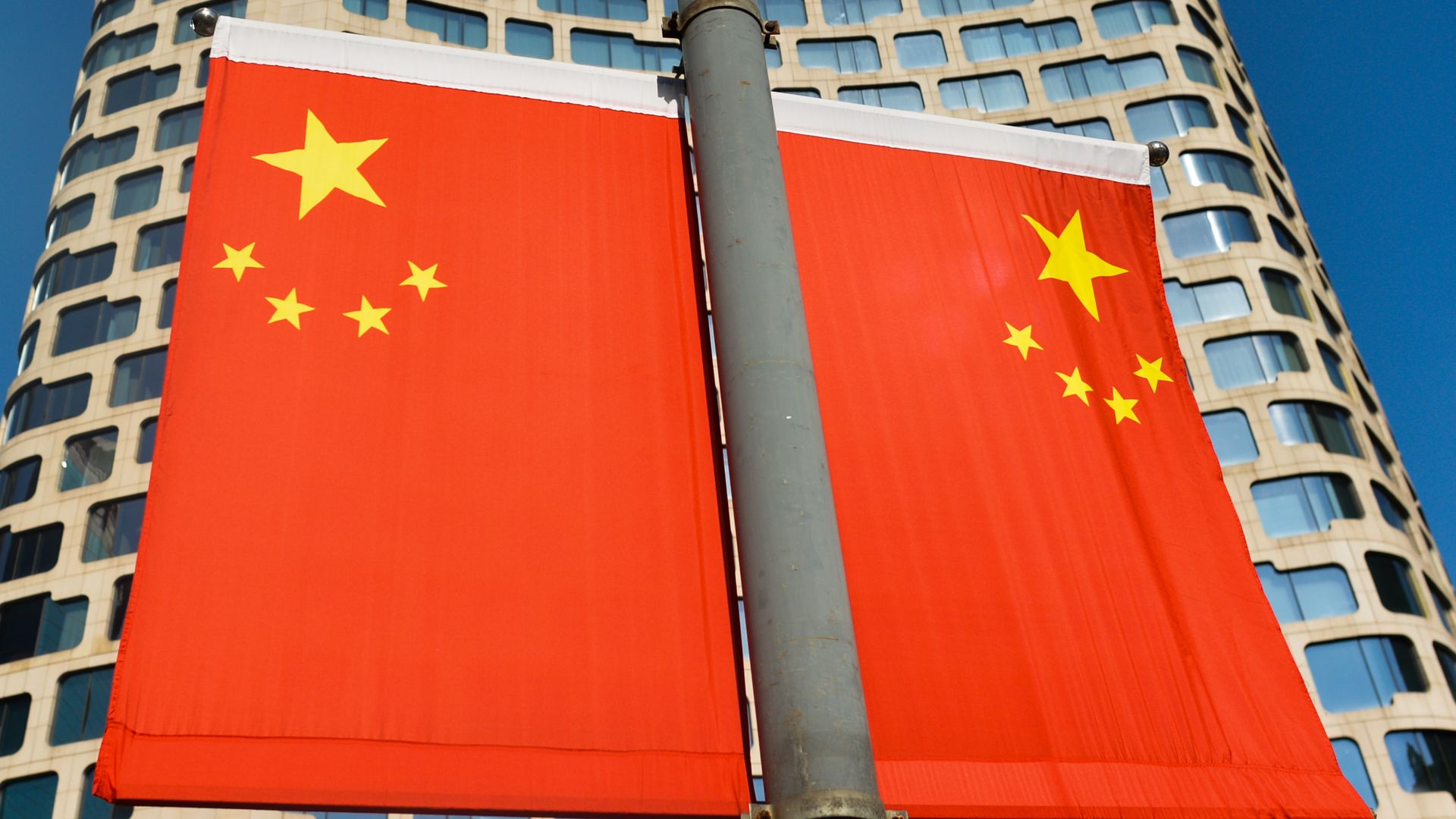Two Chinese flags on a pole before a building with lots of windows.