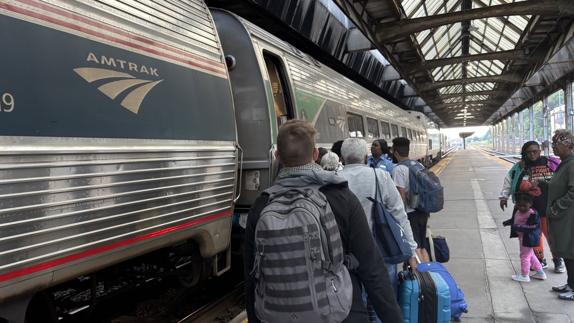 People with luggage boarding a silver Amtrak train at a covered railway platform with glass roof panels during daytime.