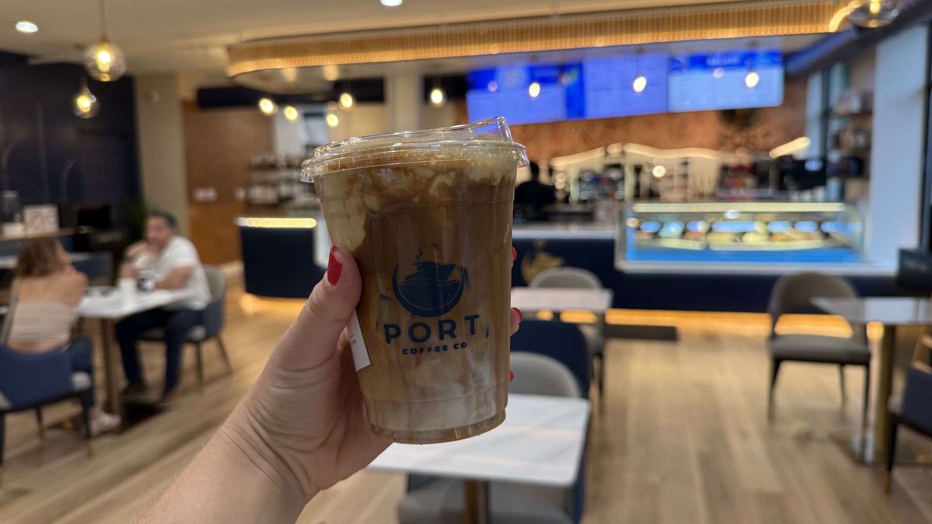 Hand with red nails holds a tall iced coffee in a clear-lid cup at a bright cafe, with warm pendant lights, blue menu boards, and wooden floors in the background.