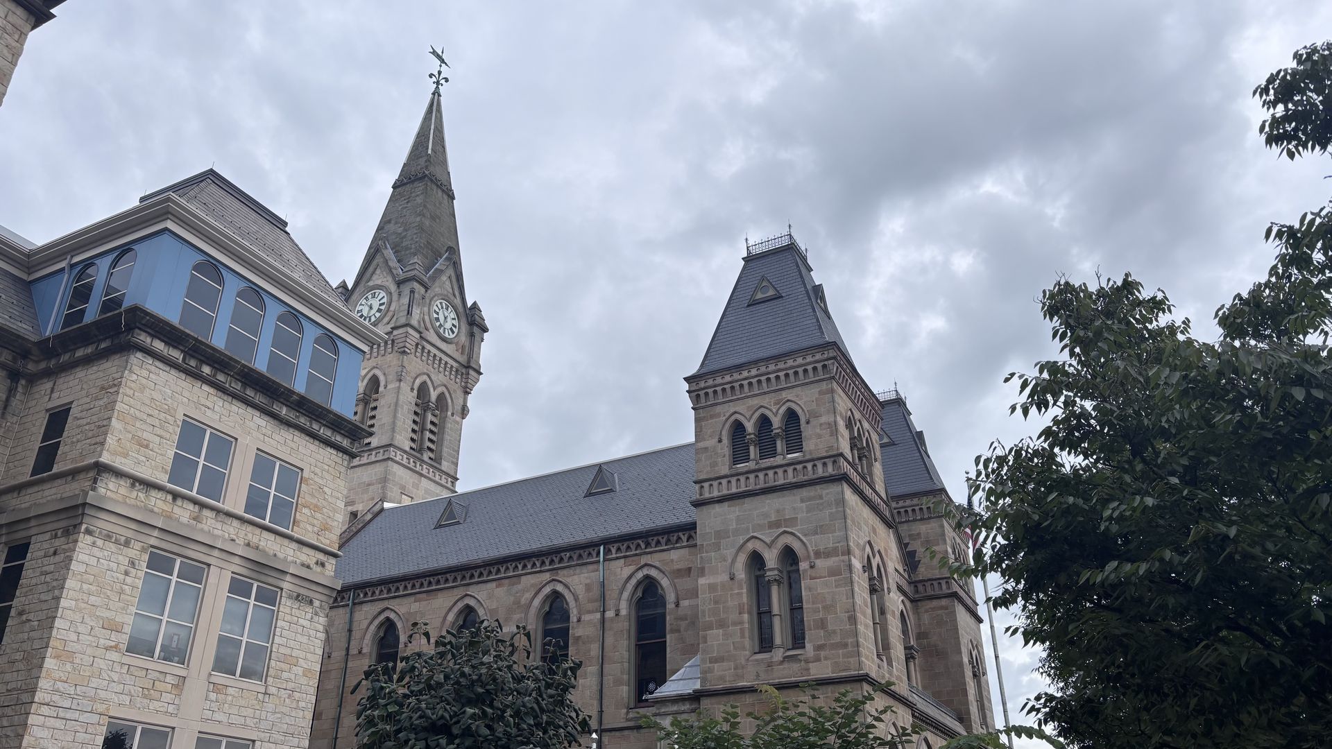Historic stone building with two clock towers and a tall spire, blue-trimmed arched windows on the left, gray slate roofs, and an overcast sky framed by trees.