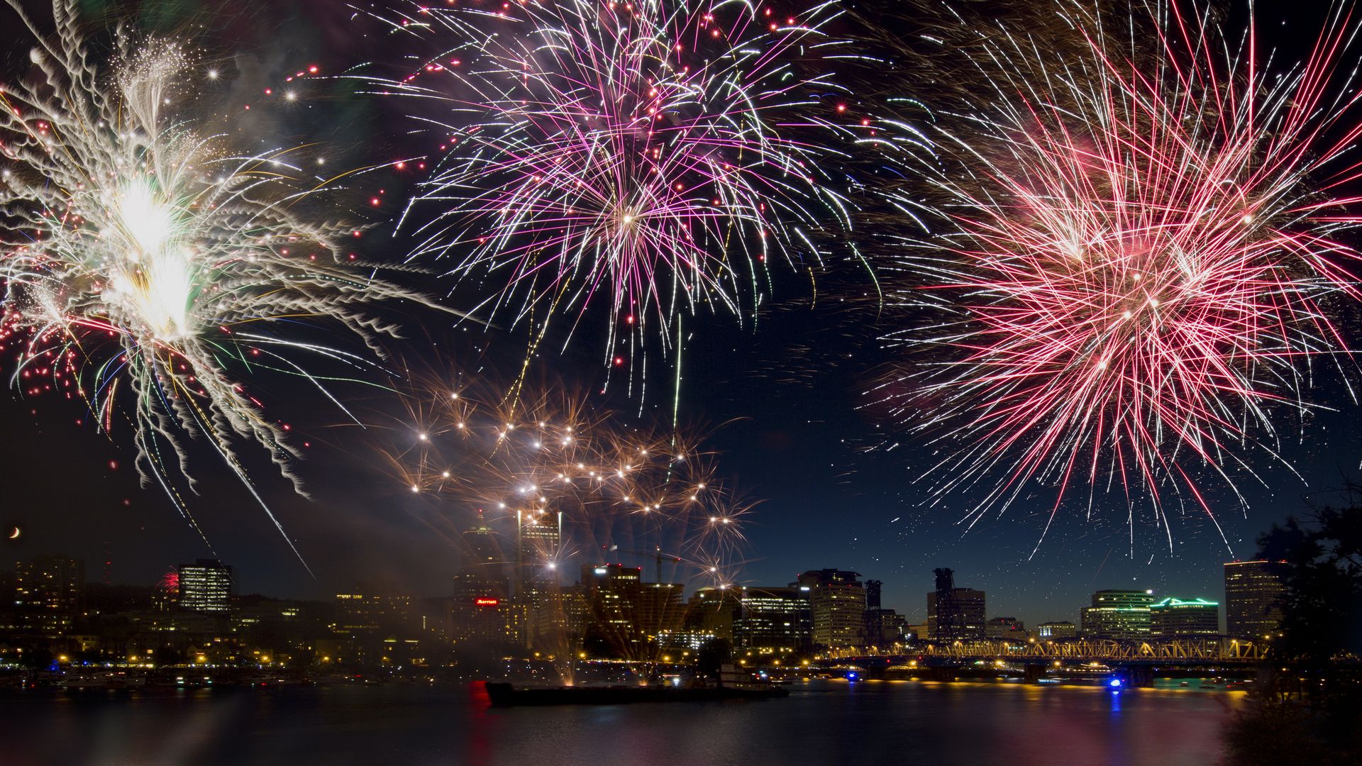 Nighttime city skyline beside a dark river as multicolored fireworks explode across the sky. Pink, white, red, and gold bursts illuminate tall buildings, a bridge, and the water below with reflections.