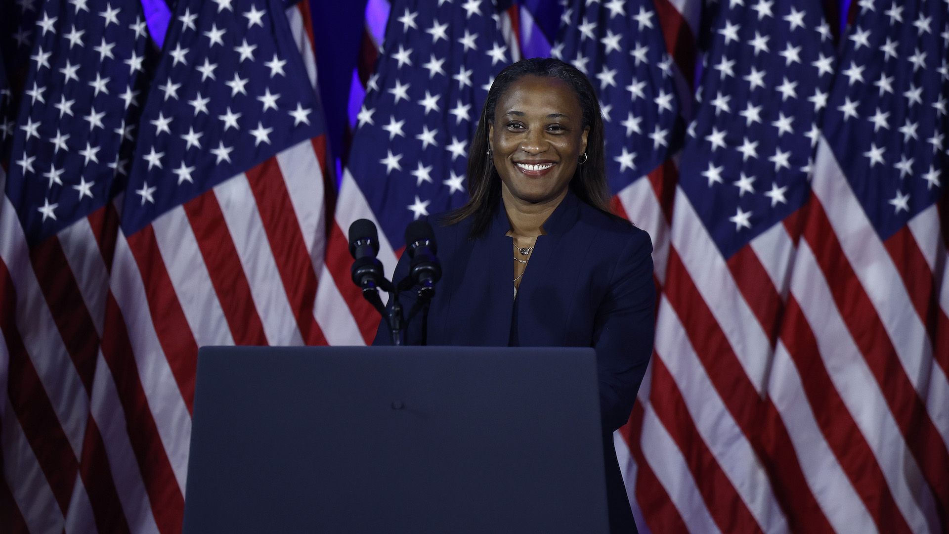 A Black woman in a navy suit stands at a podium smiling with American flags on stage behind her. 