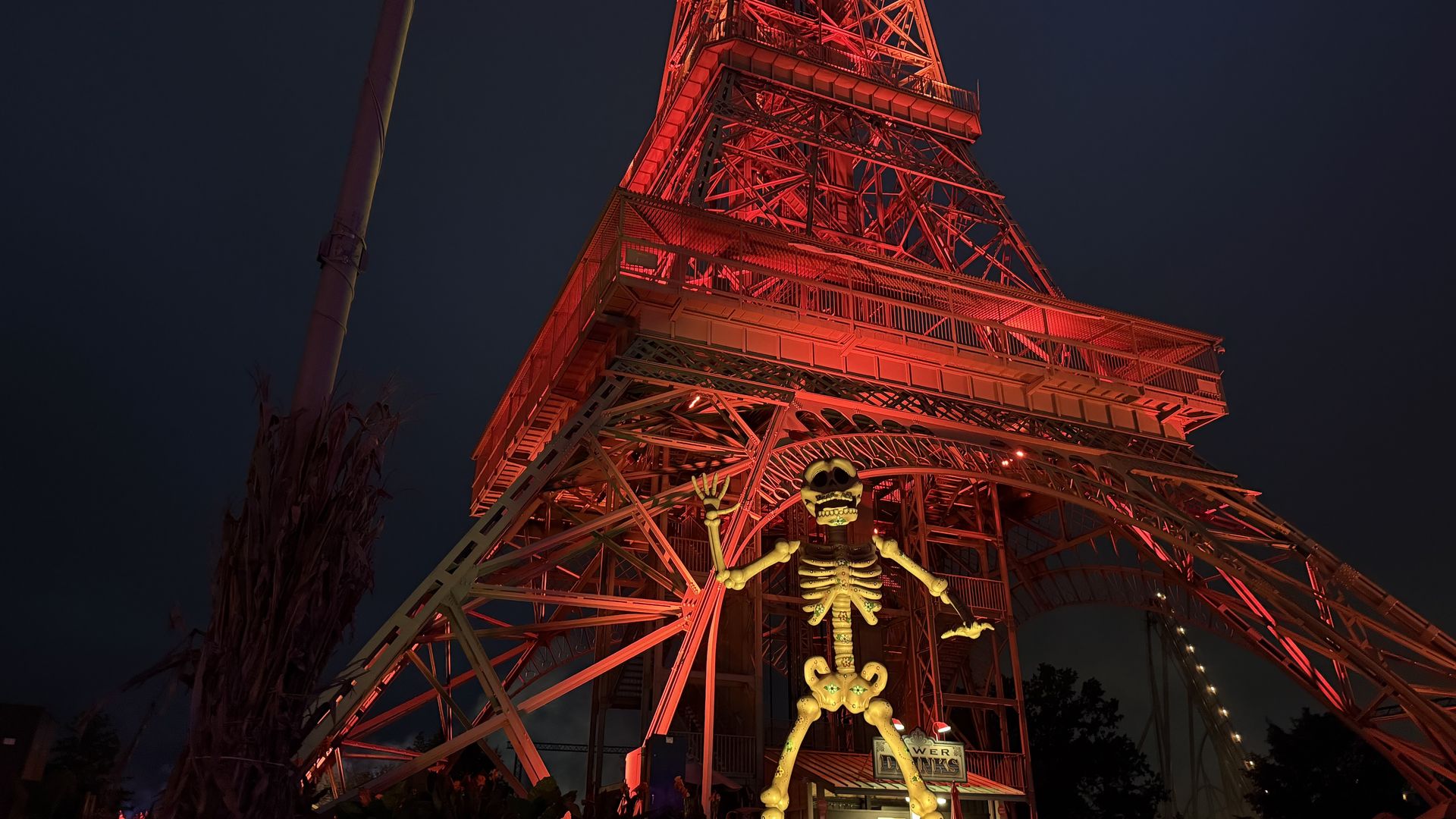 Nighttime view of a tall red-lit steel roller-coaster frame, its lattice beams glowing against a dark blue sky. Illuminated skeletal figures stand at the base, adding a spooky Halloween vibe.