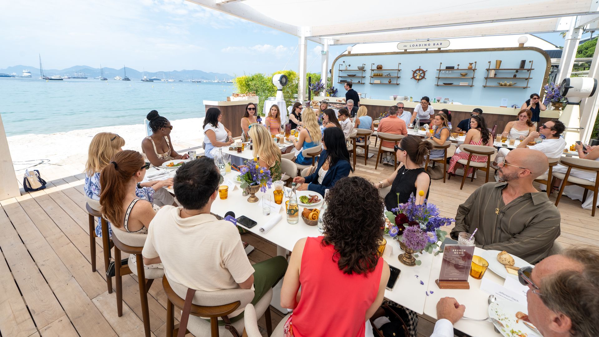 Outdoor beachside brunch with dozens of guests at long white tables on a wooden deck, sea and boats in the background, and a blue wall with shelves behind labeled "LOADING BAR".