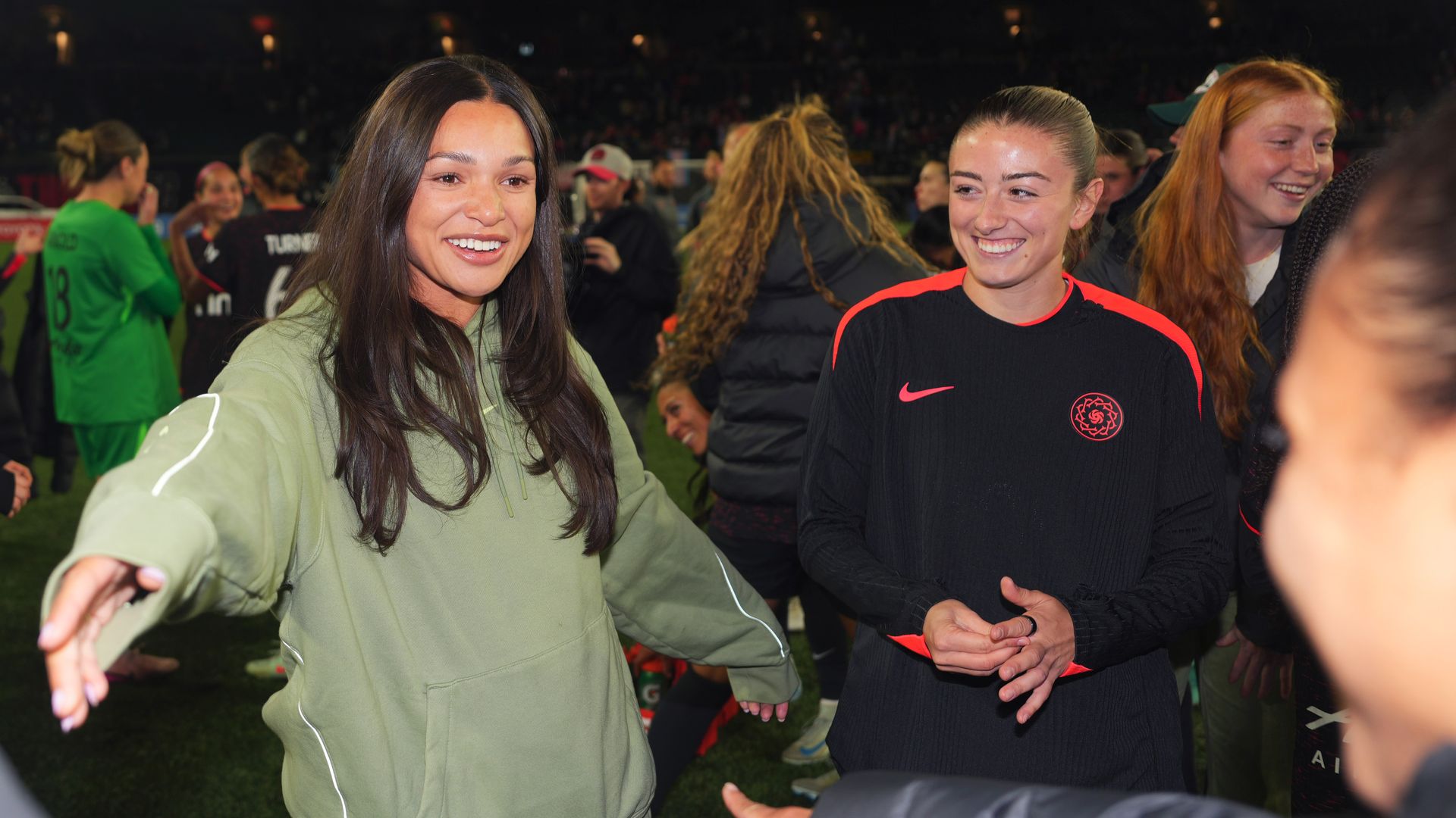 Two women smiling and interacting on a sports field at night; one in a green hoodie reaching out, the other in black and red sportswear clapping, with others in the background.