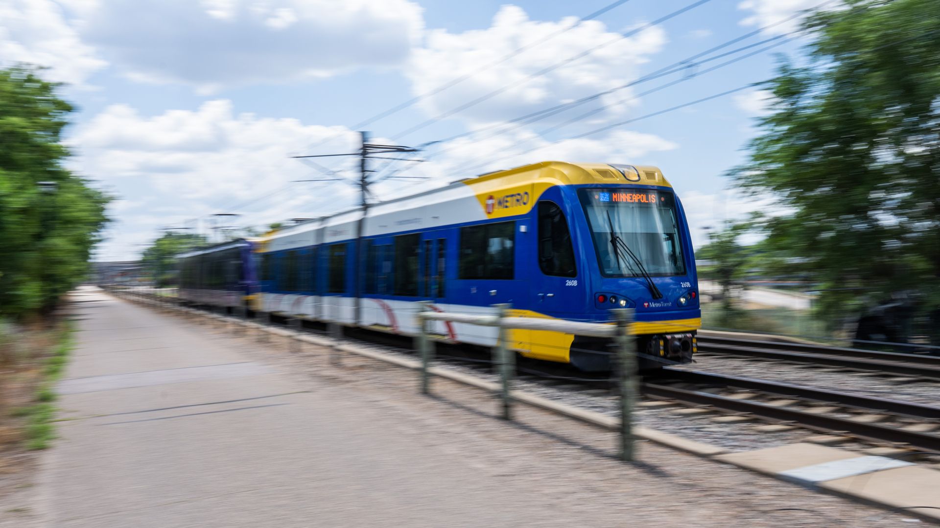 Blurry blue and yellow metro train on tracks, moving past green trees under a bright blue sky with white clouds; overhead power lines stretch across the scene.