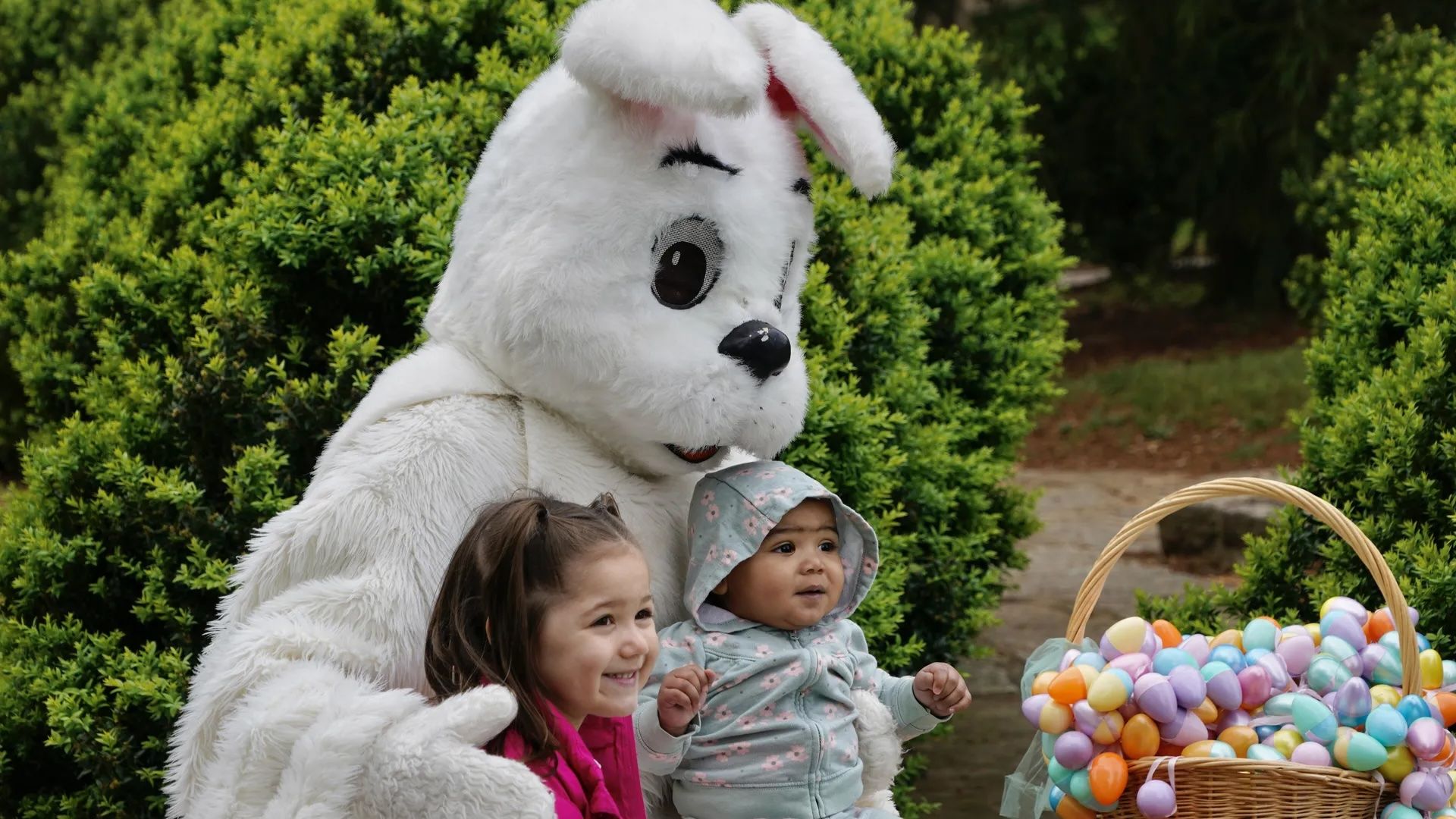 A person in a fluffy white Easter bunny costume stands with two young children outdoors, beside a wicker basket filled with colorful pastel Easter eggs, with green bushes in the background.