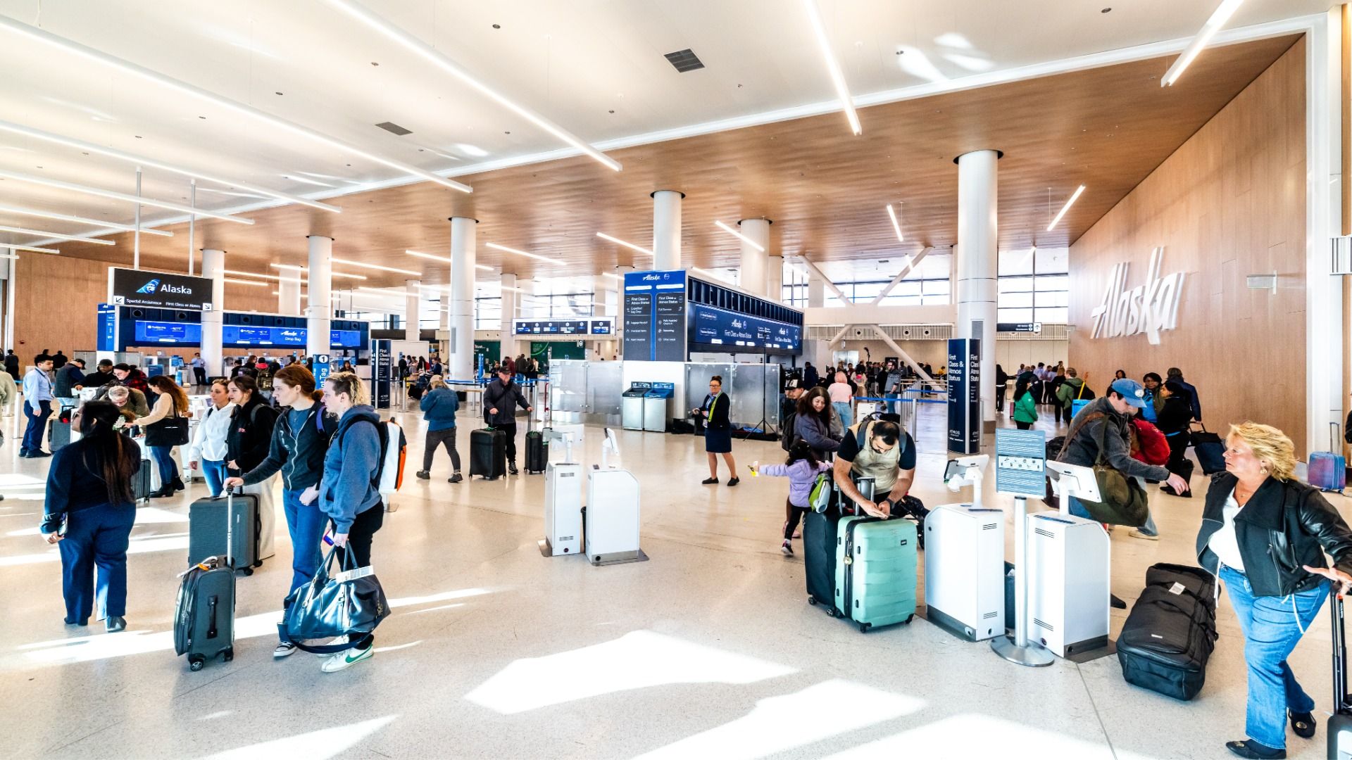 Busy airport check-in area with Alaska Airlines sign on wood-paneled wall. Passengers line with rolling suitcases, blue information signs, counters, and bright linear lights overhead.