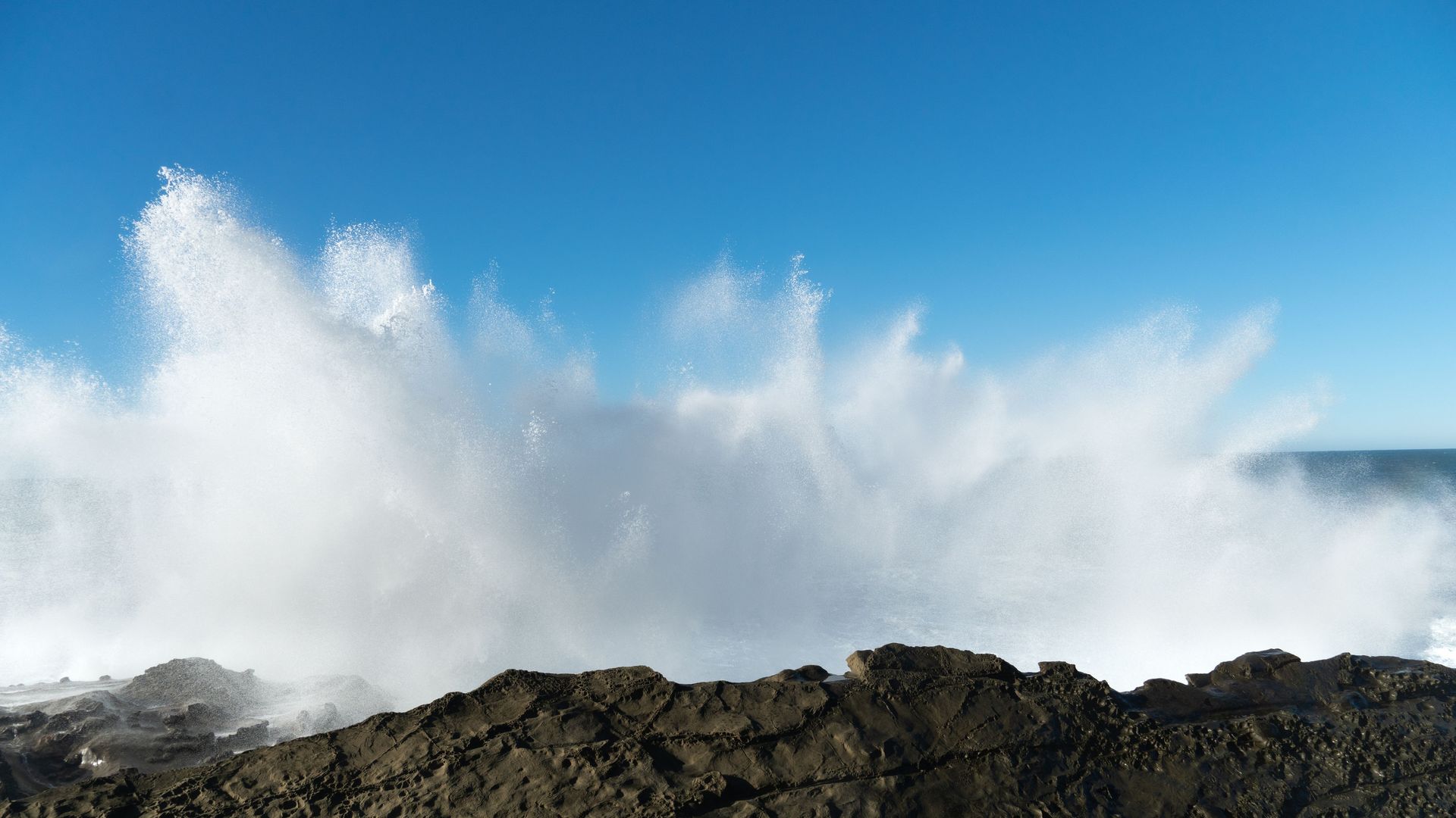 Large white sea spray burst against clear blue sky, crashing over dark rugged rocks along a coastline.
