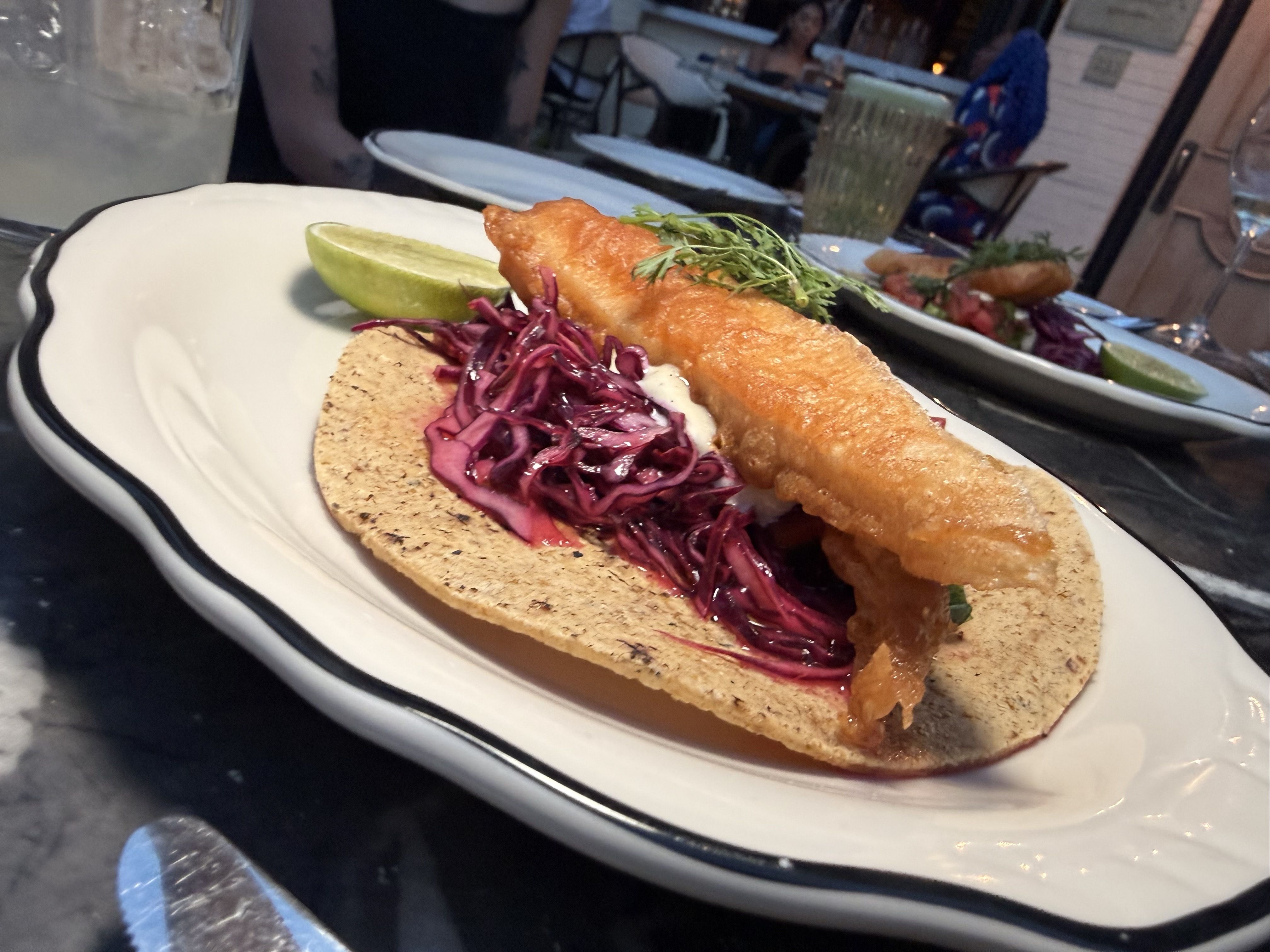 Crispy fried fish taco on a corn tortilla with purple cabbage slaw, lime wedge, and fresh herbs; plate in a restaurant with another dish and glasses blurred in the background.