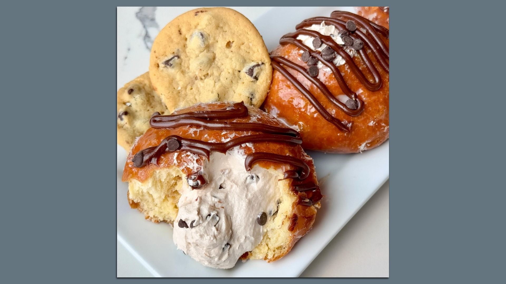 Close-up of two glazed doughnuts topped with chocolate drizzle and chocolate chips, one split open showing creamy filling with chocolate chips, accompanied by two chocolate chip cookies on a white plate.