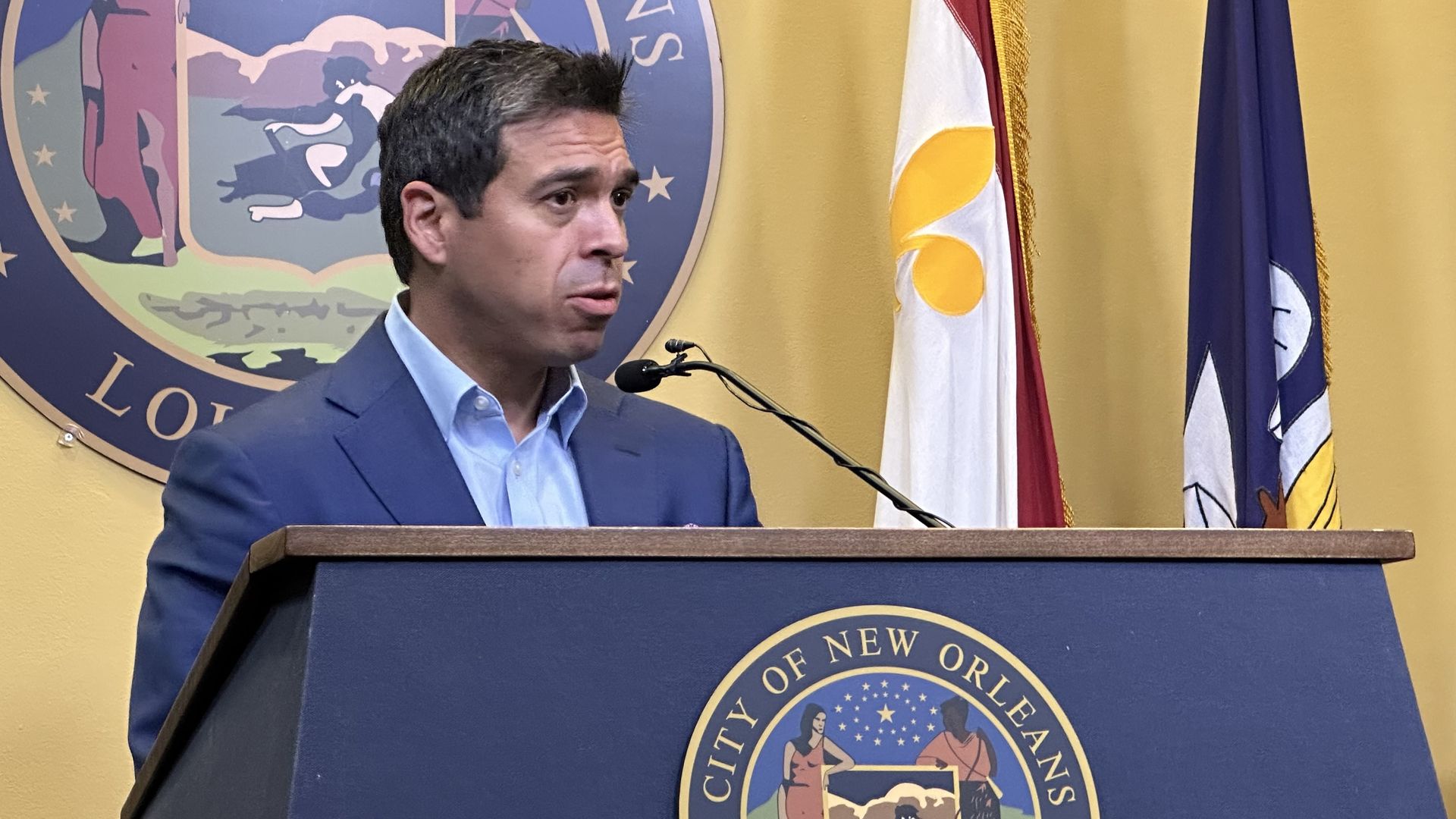 Man in blue suit speaking at a City of New Orleans podium with a microphone, with city seal and flags in the background against a yellow wall.