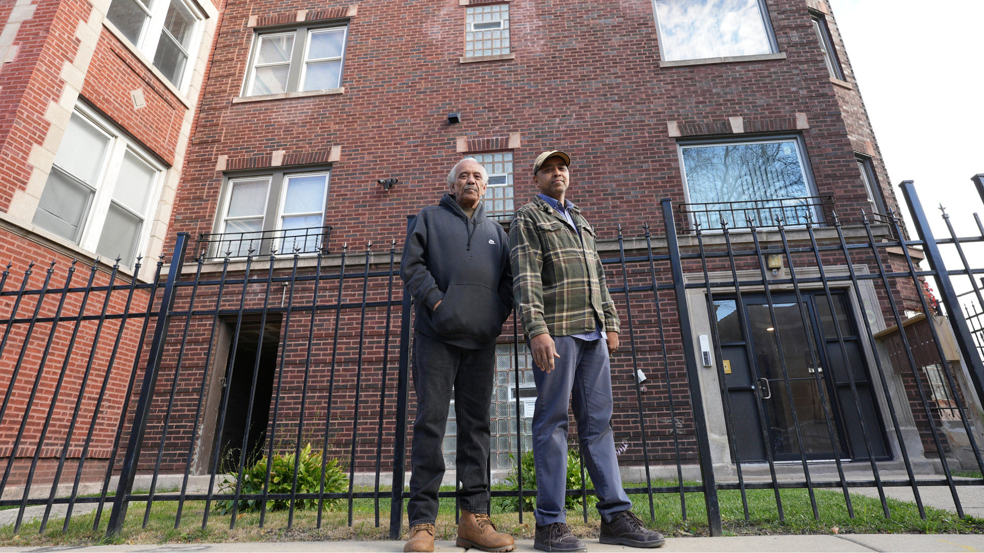Two men in a front of a brick building. 