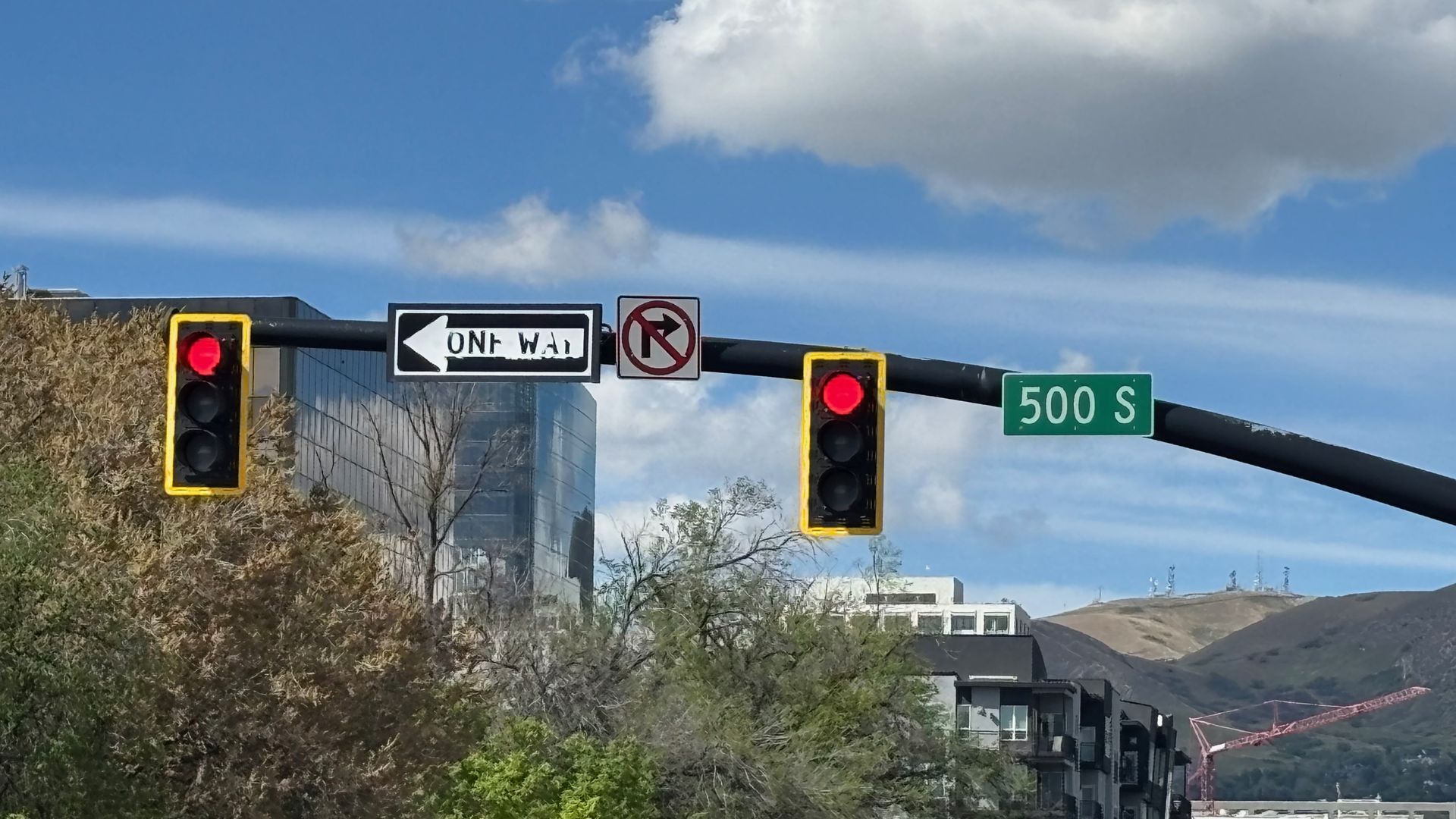 Urban intersection with two yellow-framed red traffic lights on a black overhead bar. A left-pointing "ONE WAY" sign, a no U-turn sign, and a green "500 S" street sign against a blue sky and hills.