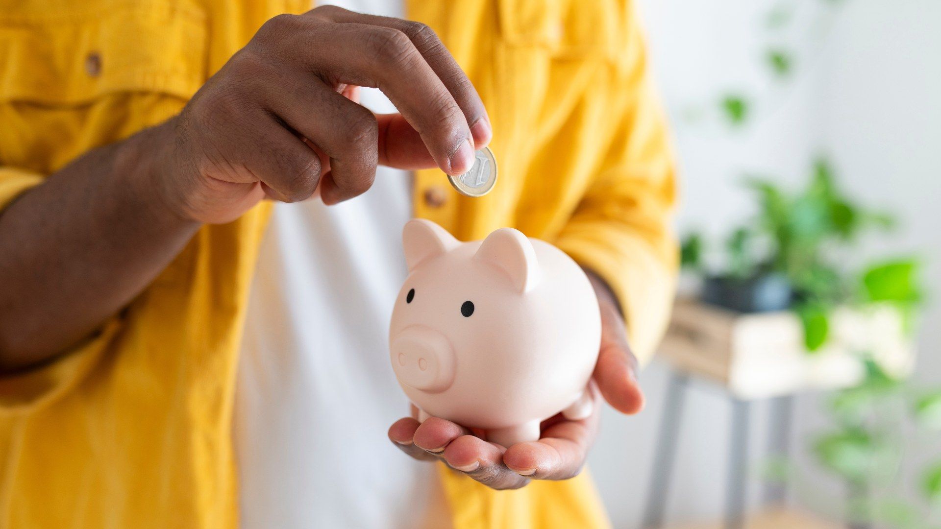 Person in yellow shirt holding a pink piggy bank and inserting a coin, with blurred green plants in the background.