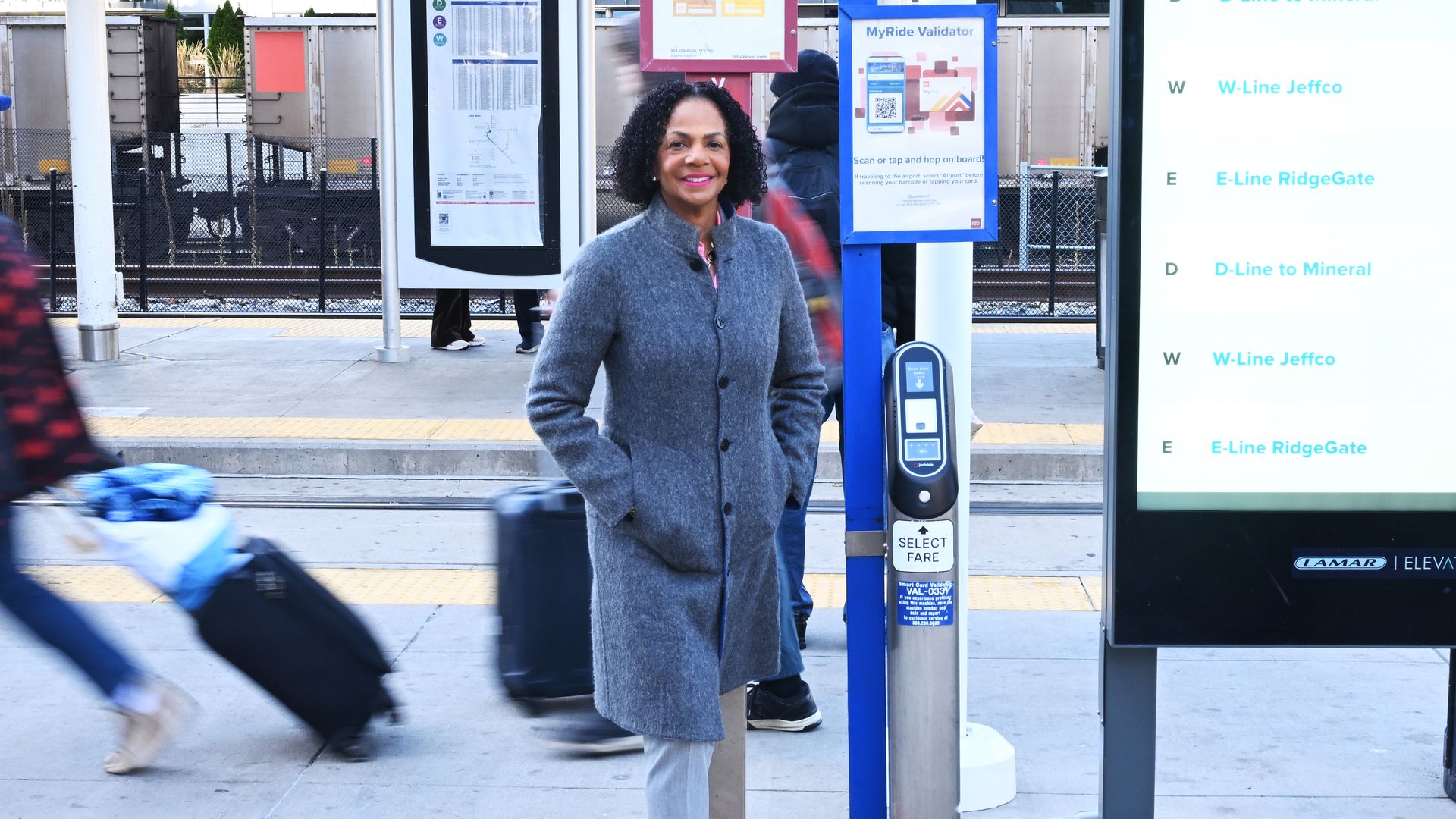 A woman in a gray coat and pink shoes stands on a train station platform beside a tall digital timetable; passing travelers with rolling luggage are in the background.