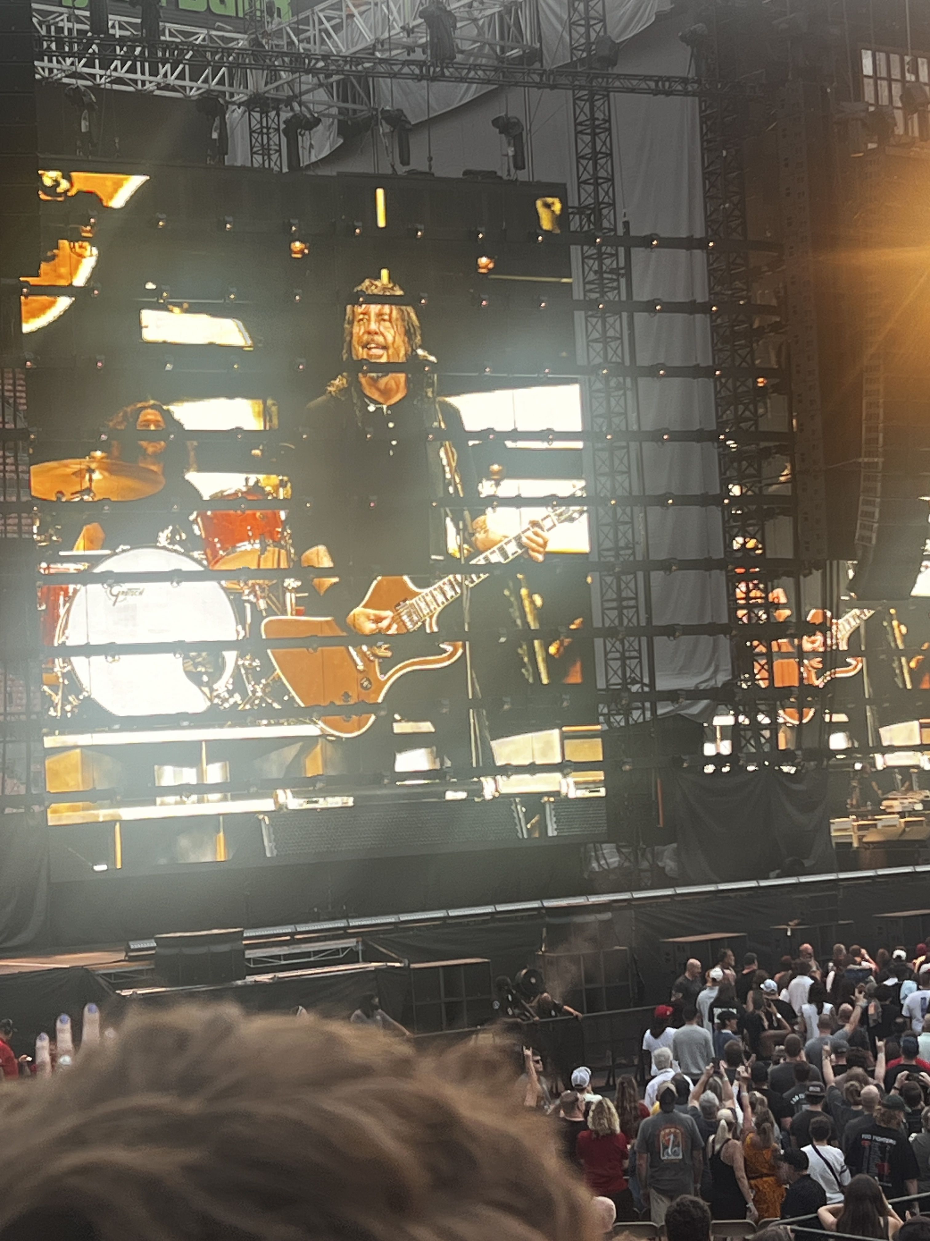 Outdoor concert stage with a guitarist center-screen, smiling while playing an electric guitar; drummer on the left, large LED panels and metal scaffolding behind; crowd in front.