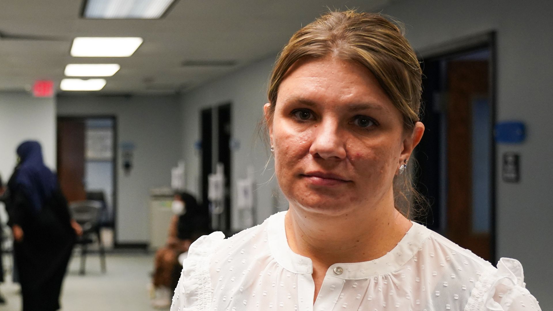 A woman in a white top poses for a portrait in a hallway of an office with indistinguishable people in the background