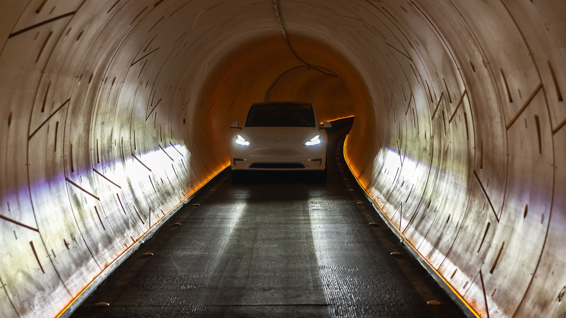 White Tesla car with headlights on driving through a lit tunnel with curved, segmented walls and orange floor lighting, branded with The Boring Company logo in top right corner.