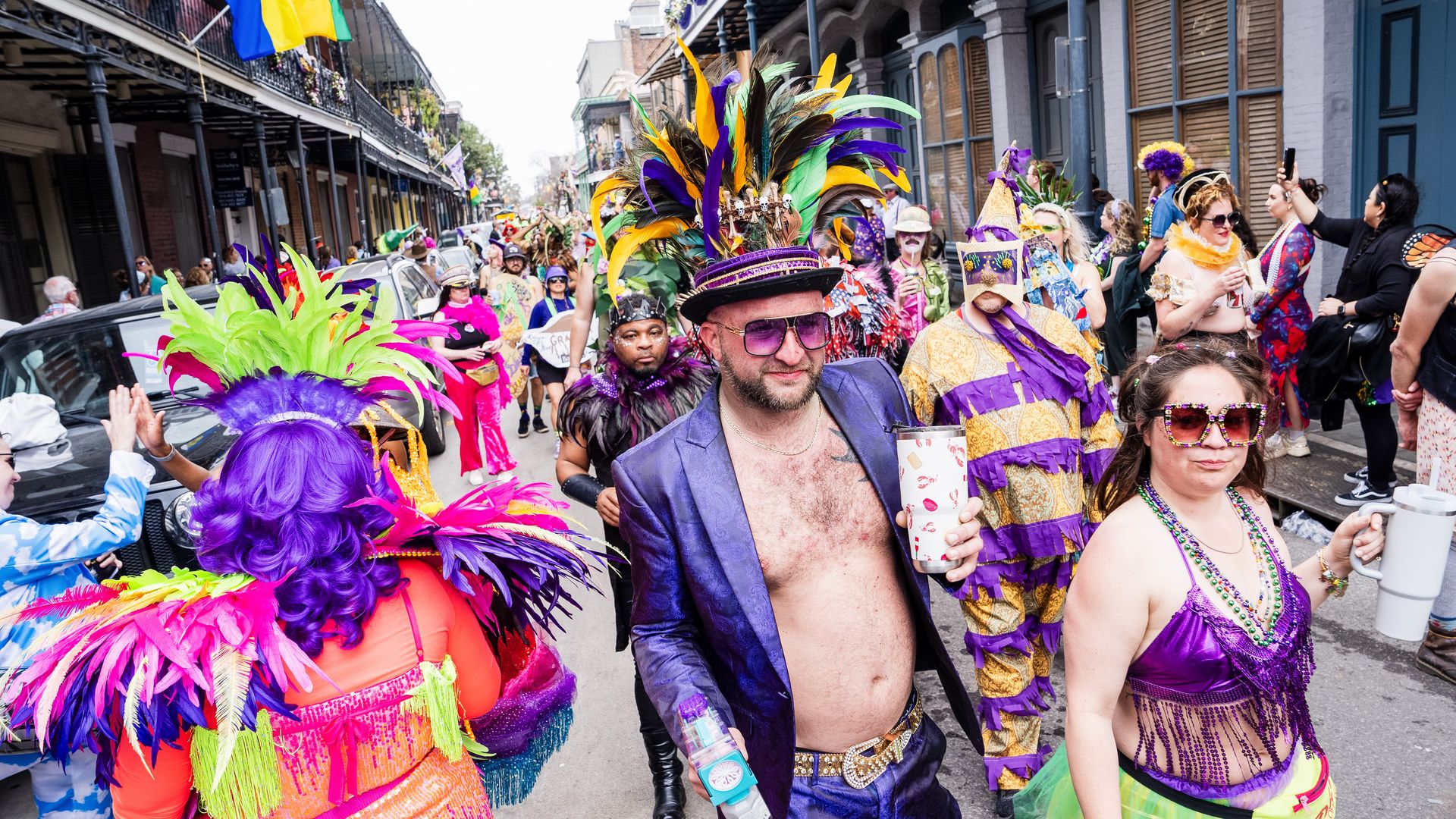 Costumed revelers fill a French Quarter street. In the foreground, a couple wears primarily purple and carries drinks as they walk.