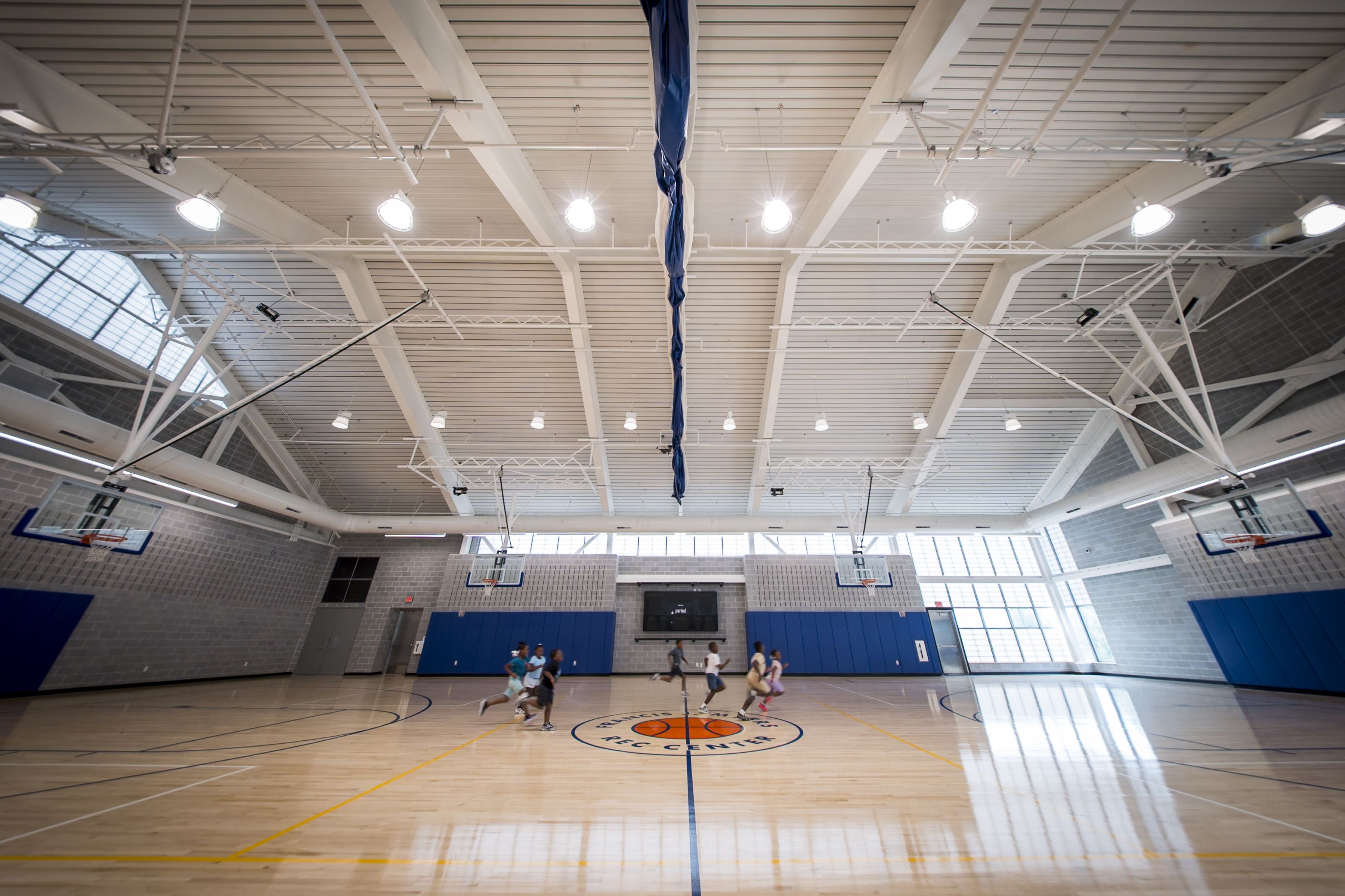 Indoor gym with a tall white ceiling and exposed beams. A blue fabric hangs from the center rafters while kids run across a polished basketball court with blue wall padding and hoops.