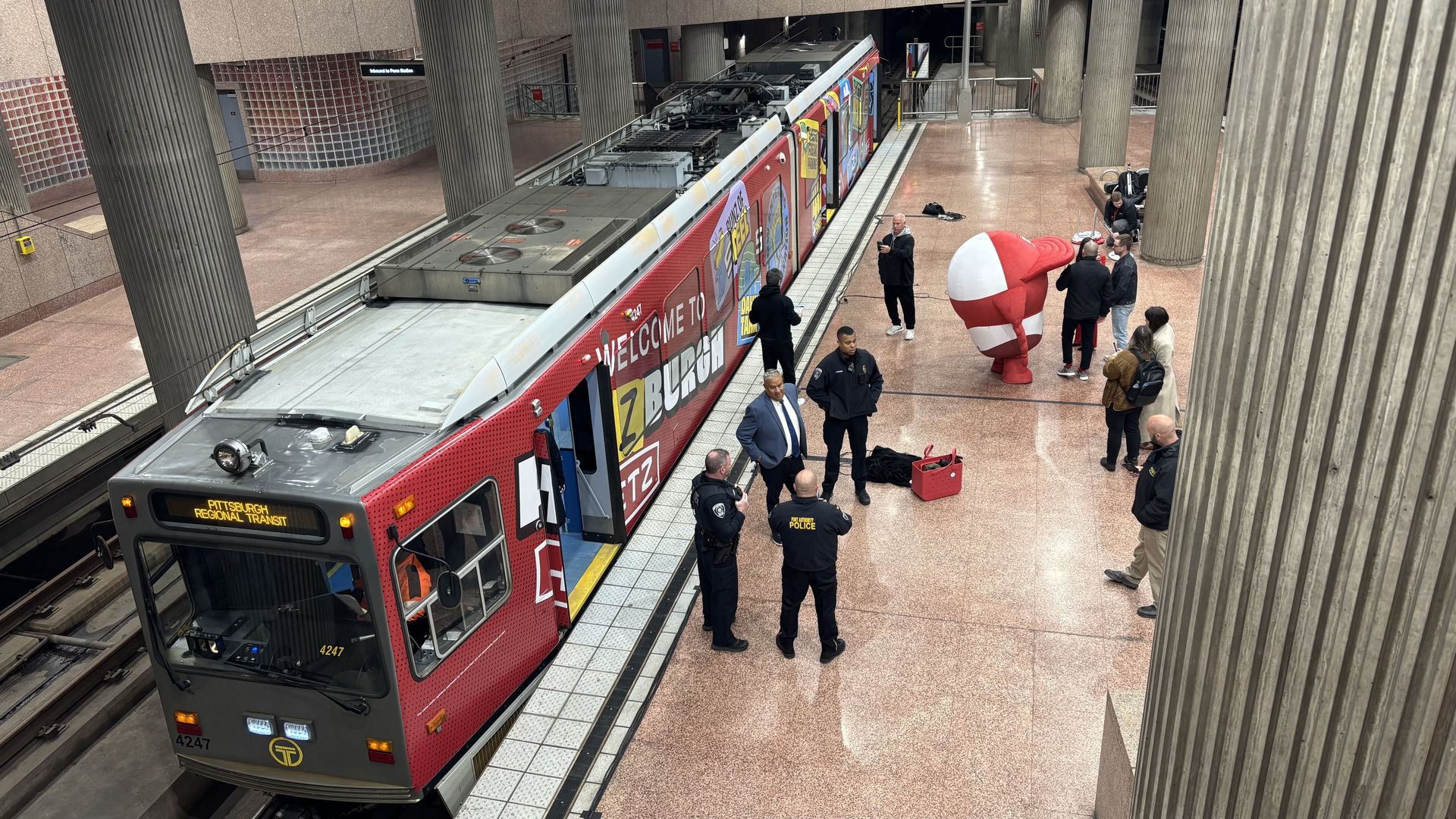 Red Pittsburgh Regional Transit train at an underground platform with tiled walls and large concrete columns; several people, including officers, stand by the doors near a large red-and-white mascot.