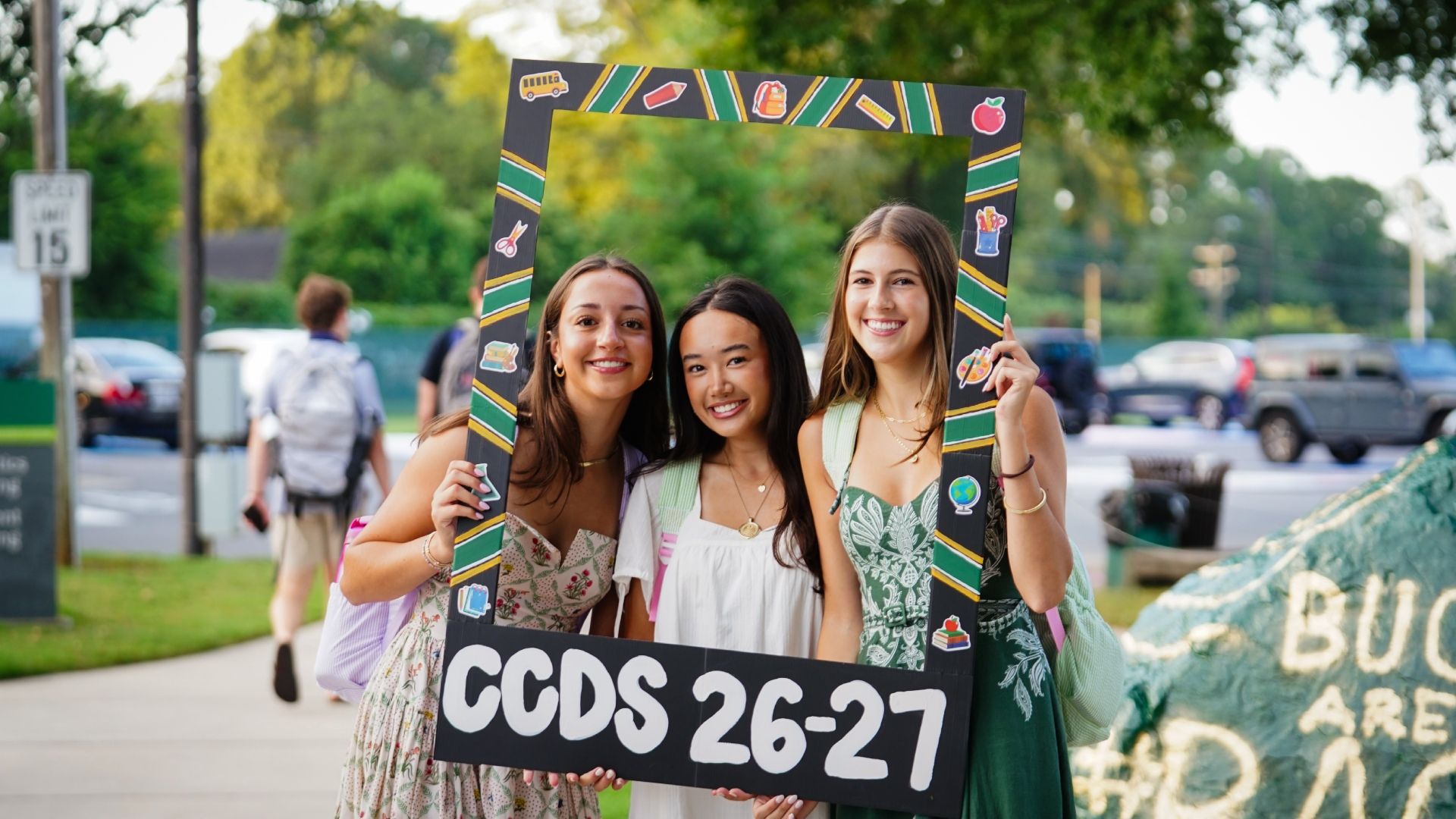 Three smiling young women pose inside a large decorated photo frame at an outdoor event; frame has school-themed stickers and reads CCDS 26-27, with trees and parked cars in the blurred background.