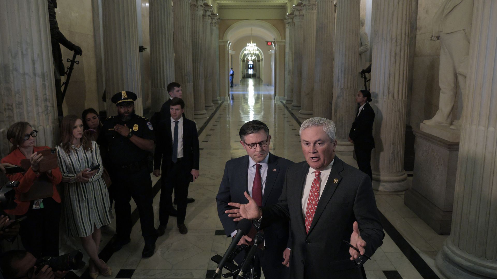 Two mean wearing suits and red ties speaking in front of a microphone in the US Capitol. The first man is speaking and has both hands out, and the other man is directly behind him watching. There are reporters and police officers in the background 