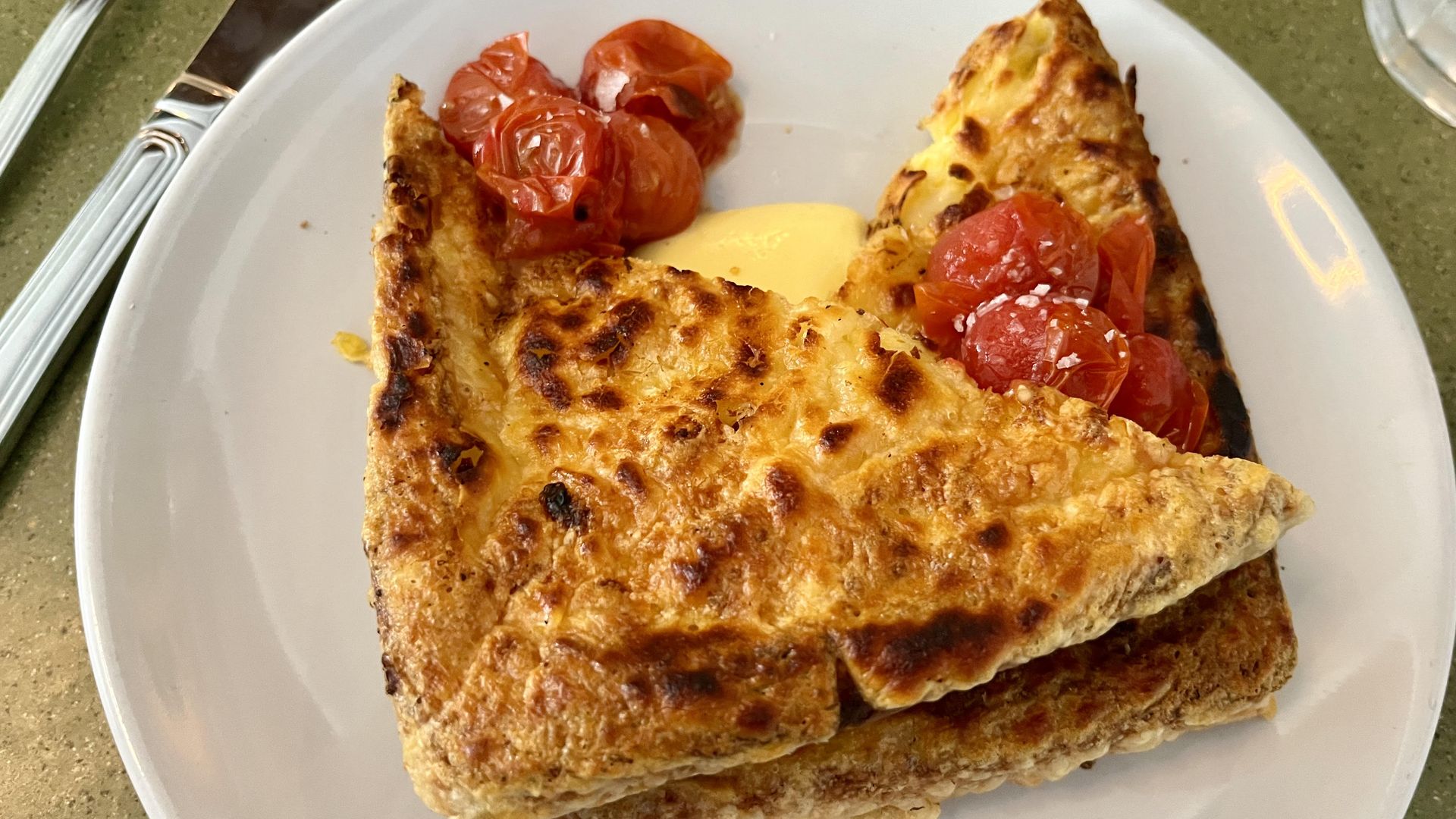 Two folded golden-brown pieces of French toast on a white plate with roasted cherry tomatoes and a dollop of yellow sauce, with a fork and knife nearby on a green table.