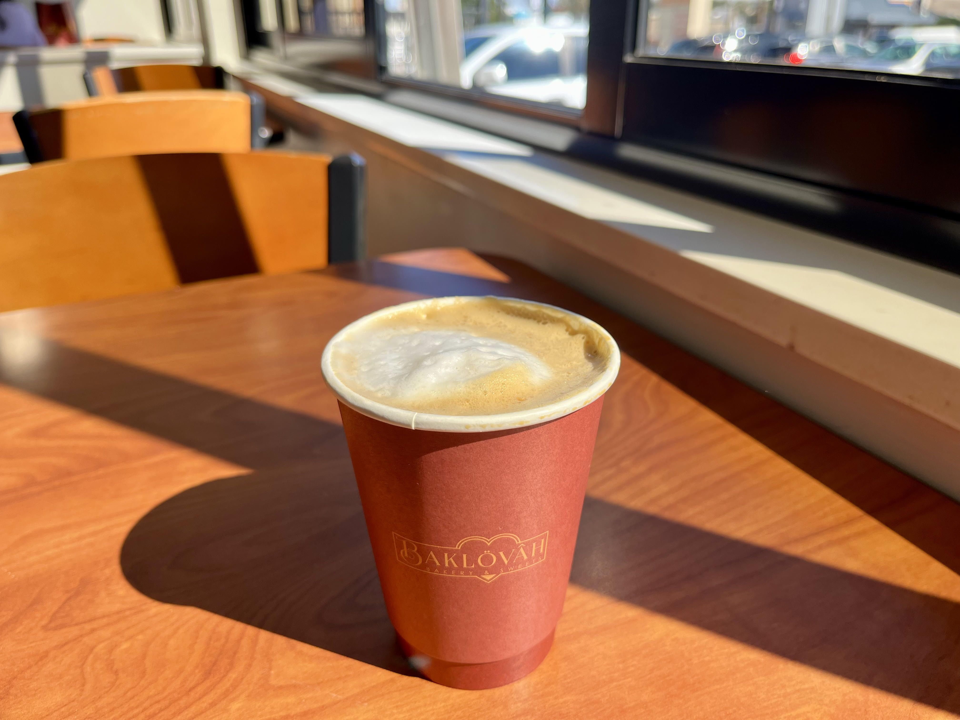 A foamy latte in a red paper cup that says "Baklovah," on a wooden table with sunlight and a shadow behind it.