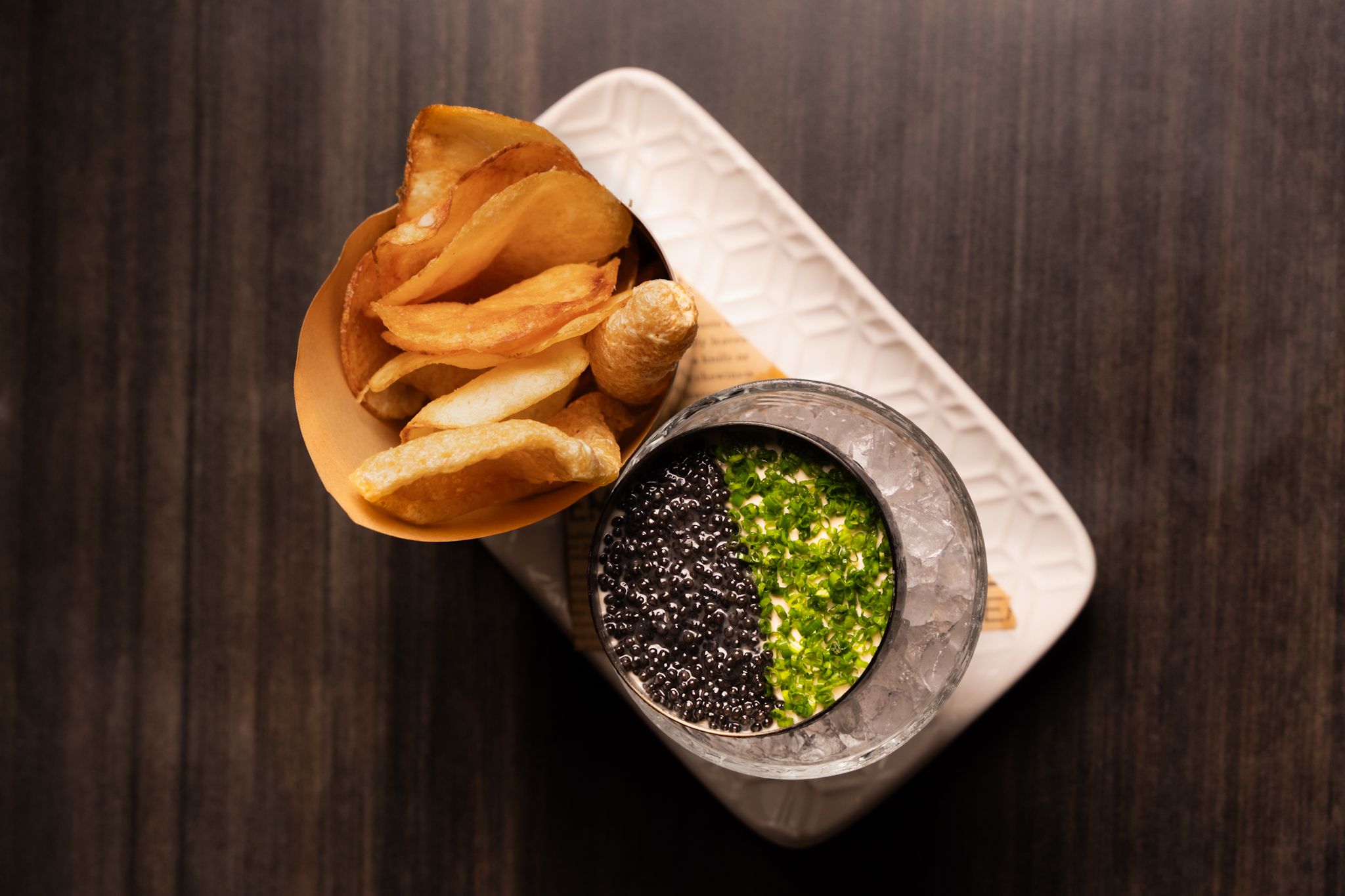 Top-down view of potato chips in a paper cone next to a black dish on ice filled with black caviar and chopped green herbs on a white rectangular plate.