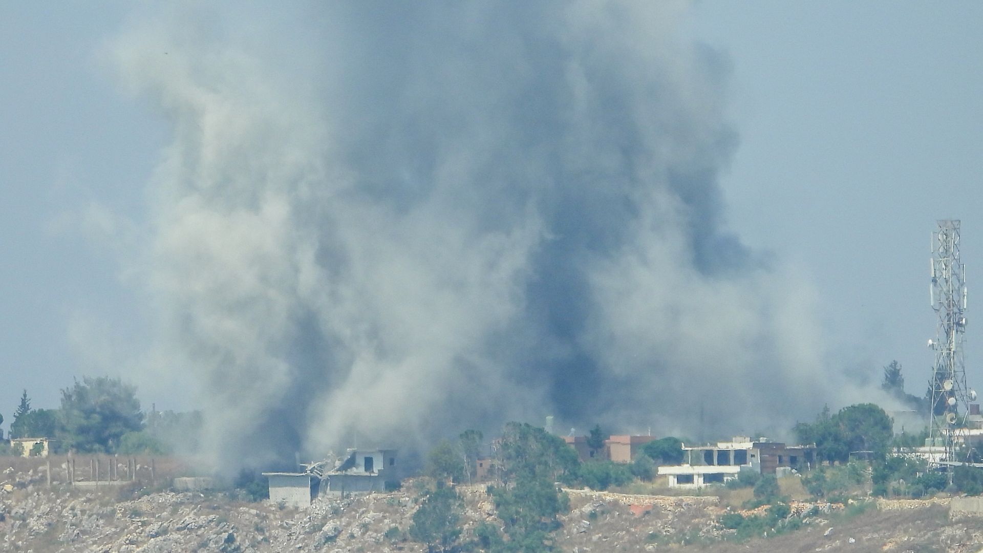 Blue sky with a large gray smoke plume rising behind a rocky field; small houses line the base and a tall cell tower stands on the right.