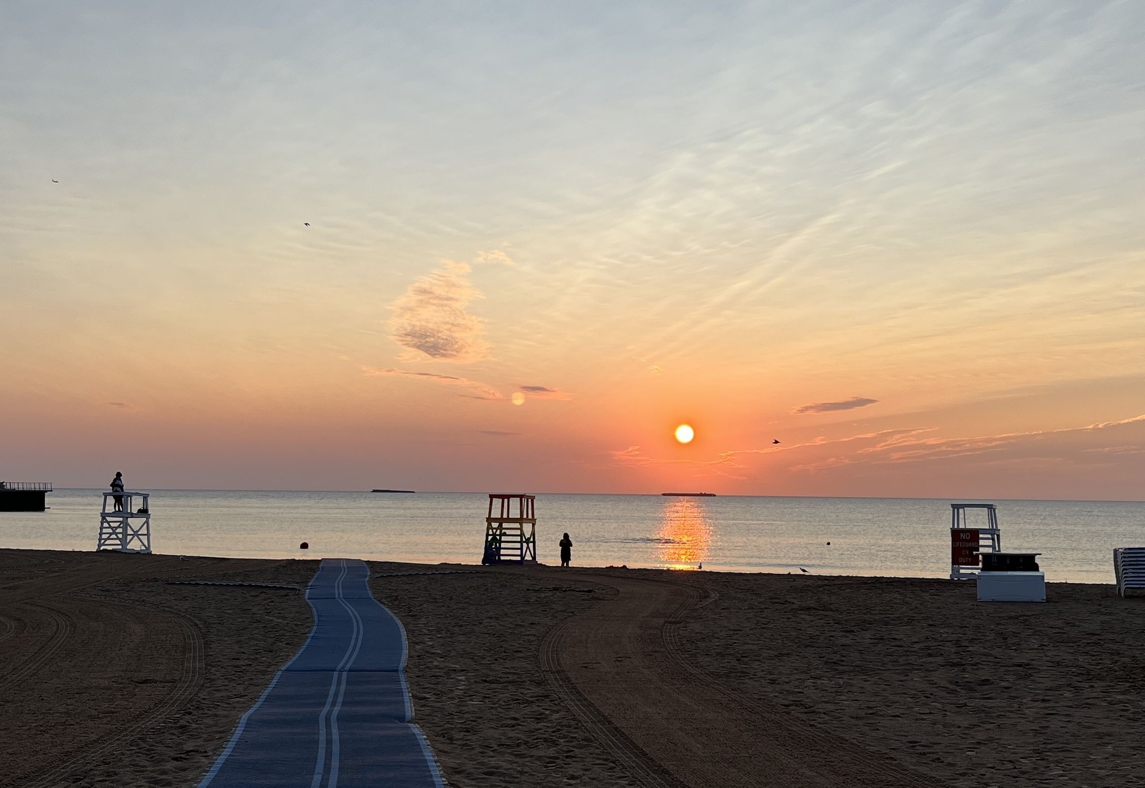 Sunset over a calm sea with an orange sun near the horizon; a blue boardwalk winds along the sandy beach, lifeguard stands on either side, a lone figure near the water.