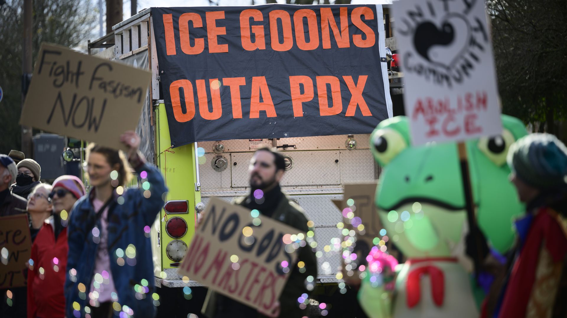 Protesters holding signs with messages like "Fight Fascism NOW" and "No Cops No Masters" stand before a vehicle with a large black banner reading "ICE GOONS OUTA PDX," one person in a frog costume.