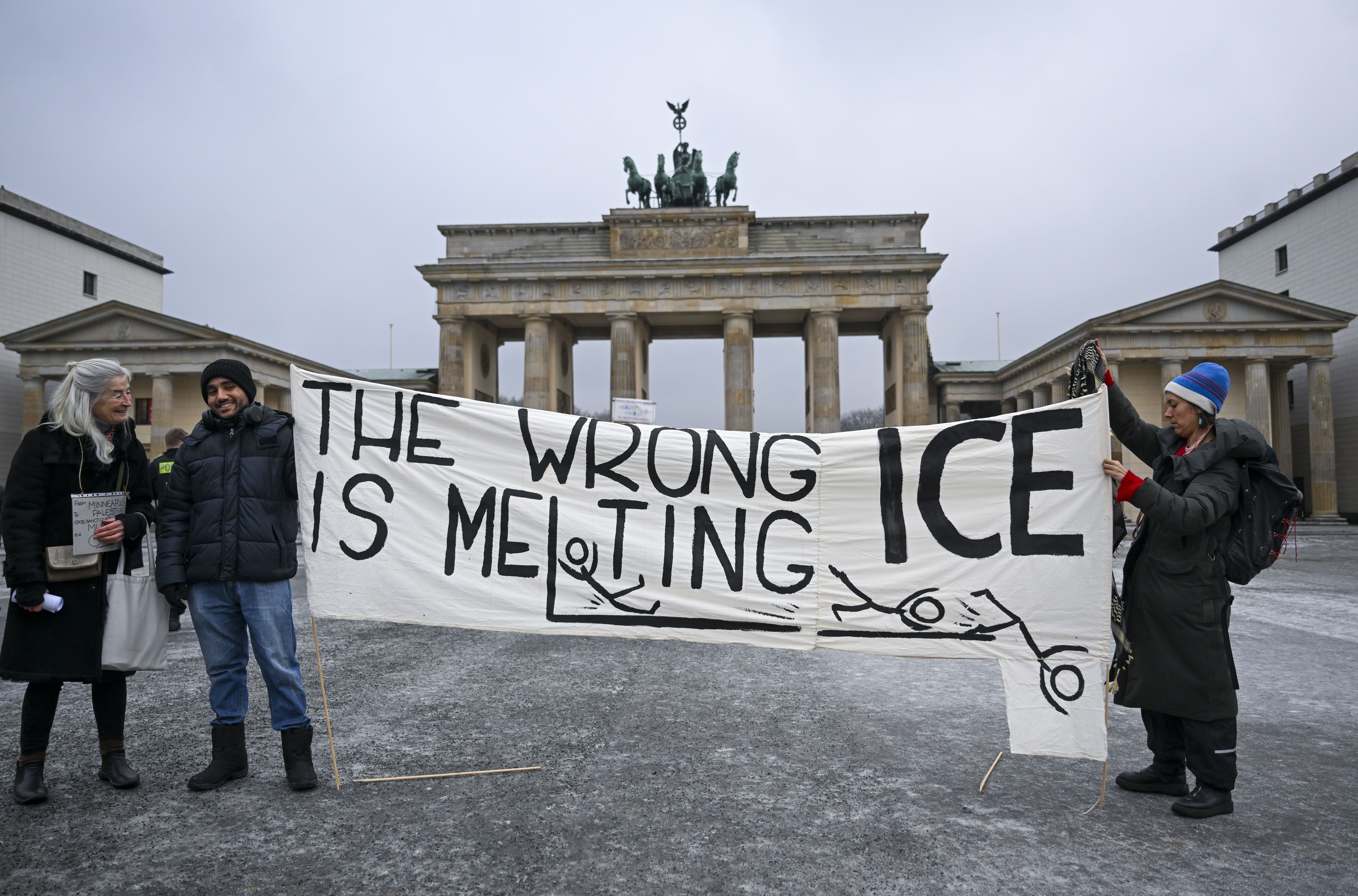 Three people holding a large white banner with black text "The wrong ICE is melting" and stick figures, standing in front of the Brandenburg Gate in cloudy weather.