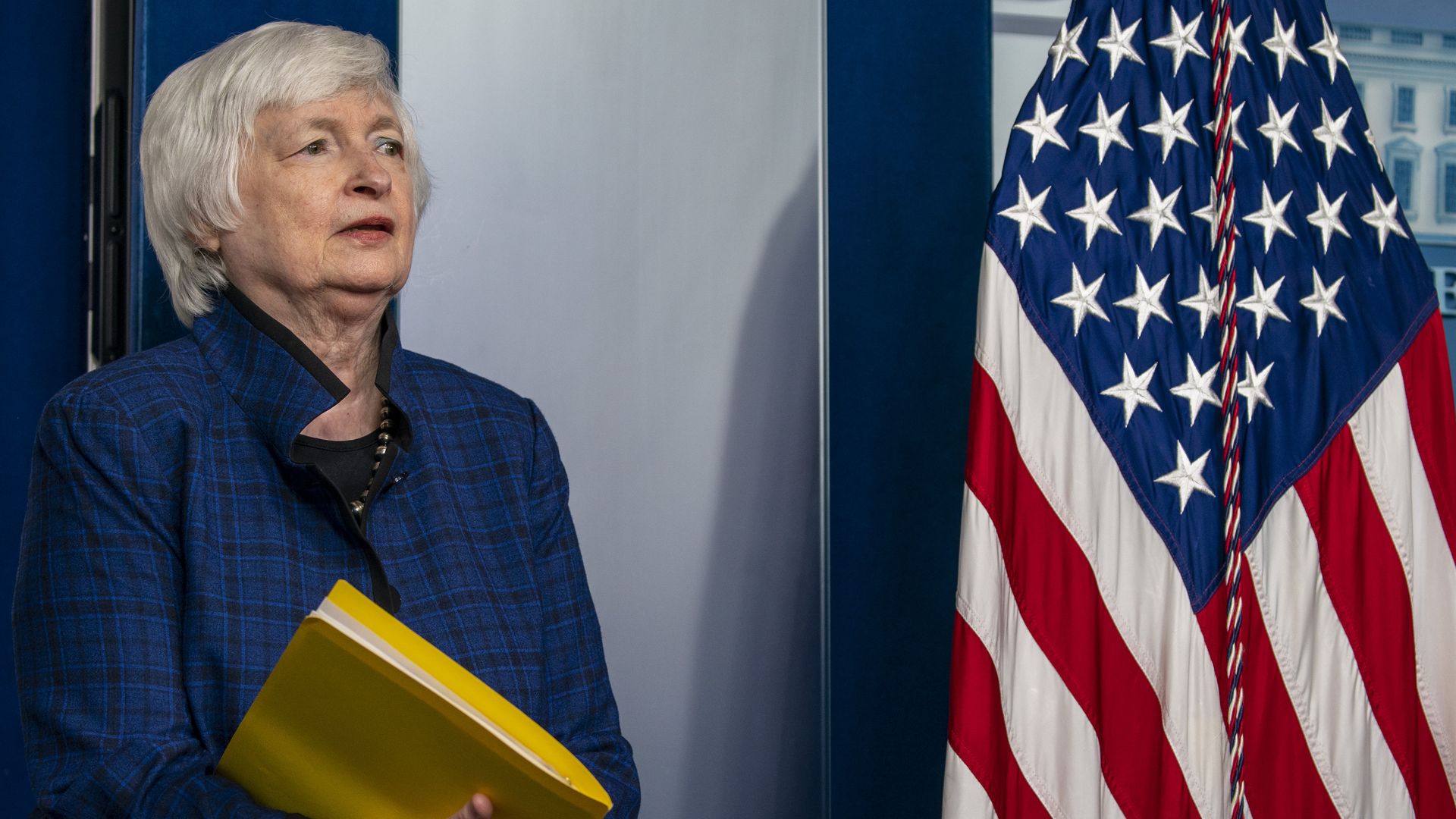 Photo of Janet Yellen holding a yellow folder and standing next to an American flag