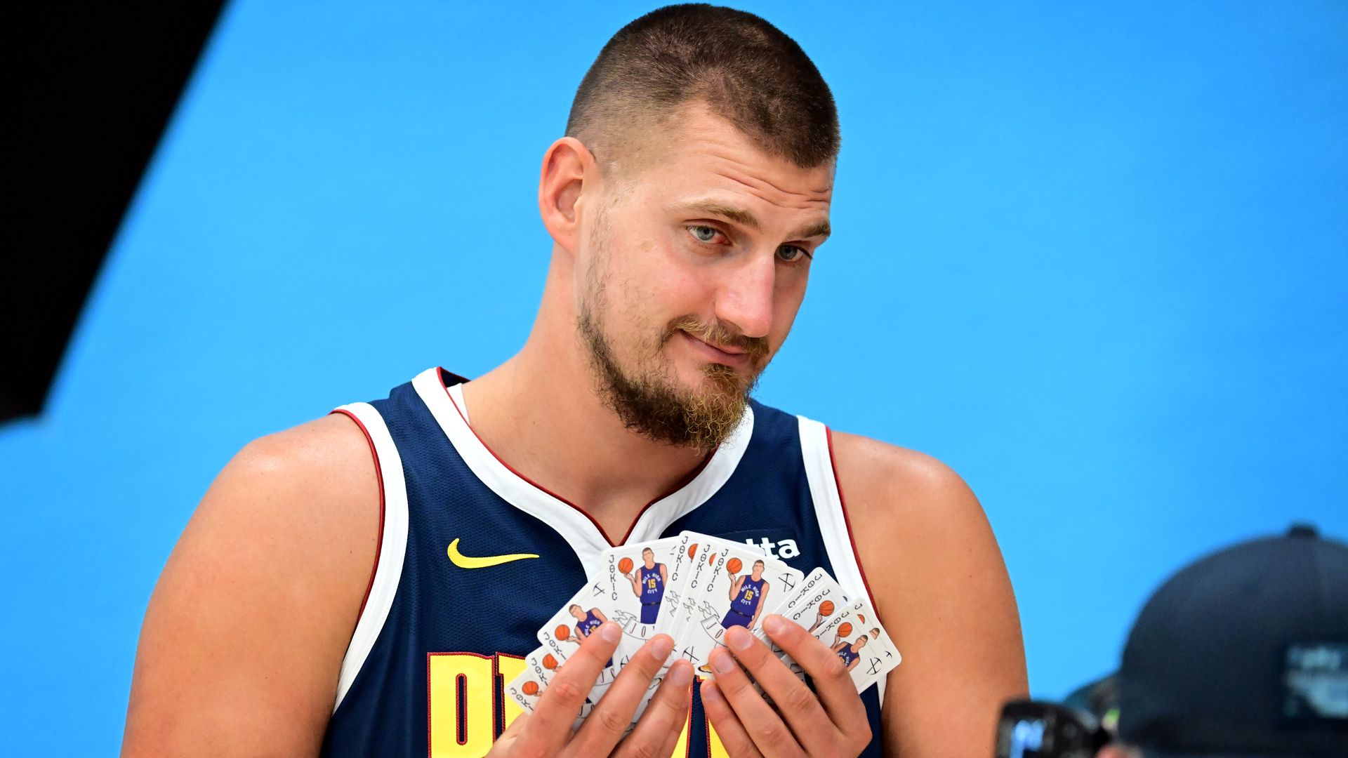 A photo of Denver Nuggets star Nikola Jokic posing for a portrait holding Joker playings cards in his hands.