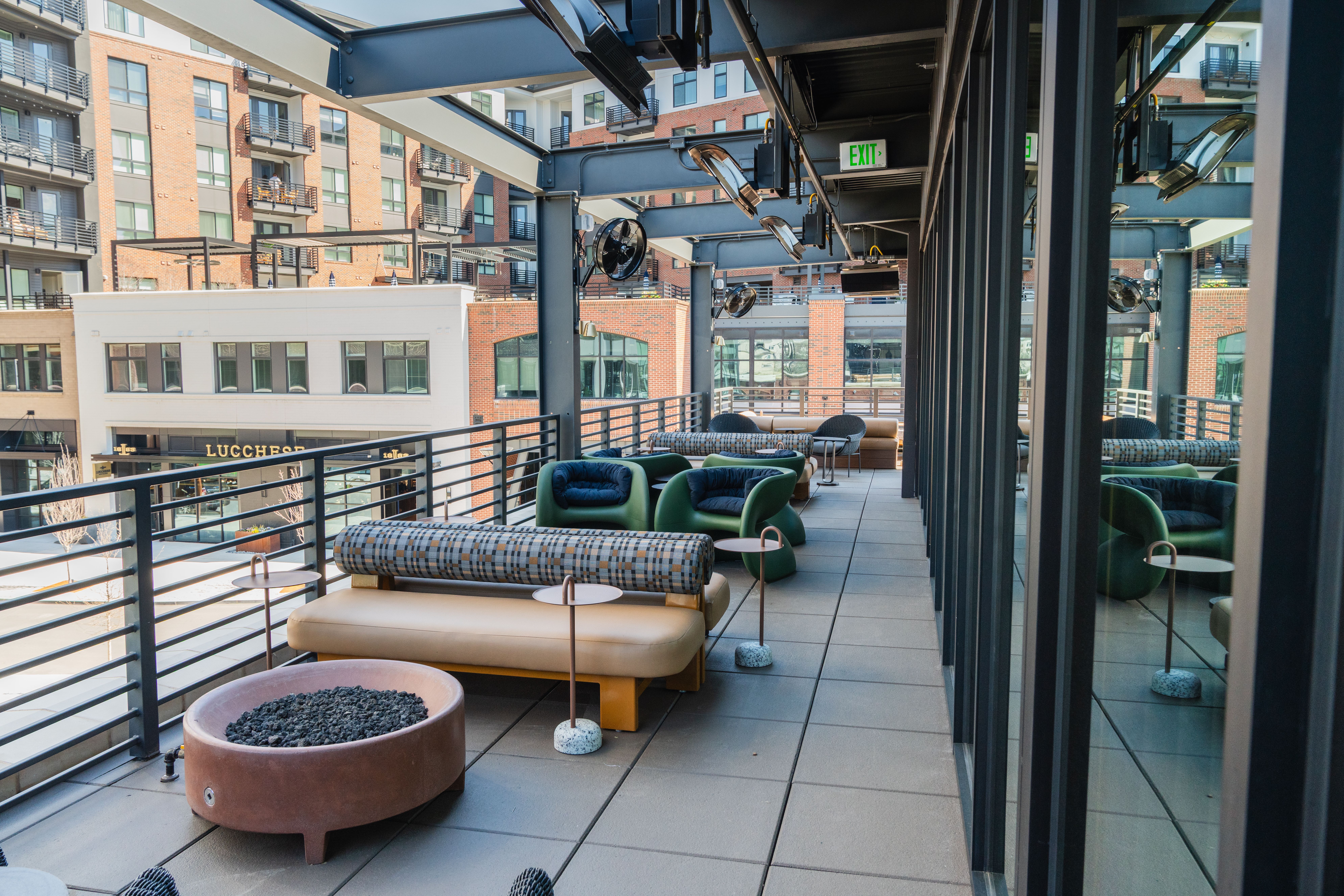 Rooftop lounge with green and patterned seating, round side tables, and a circular fire pit along a railing; urban brick buildings and glass storefronts rise in the background.