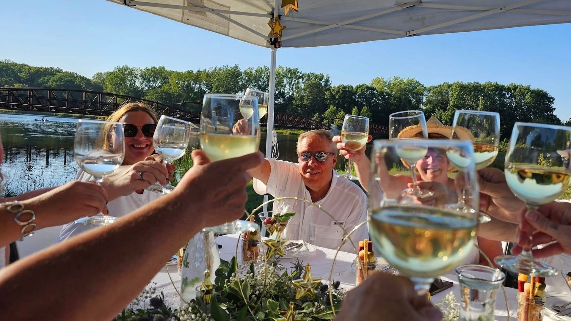 Group of friends on a sunny boat, toasting with white wine glasses under a canopy. A table with appetizers, water, a bridge and green trees visible in the background.
