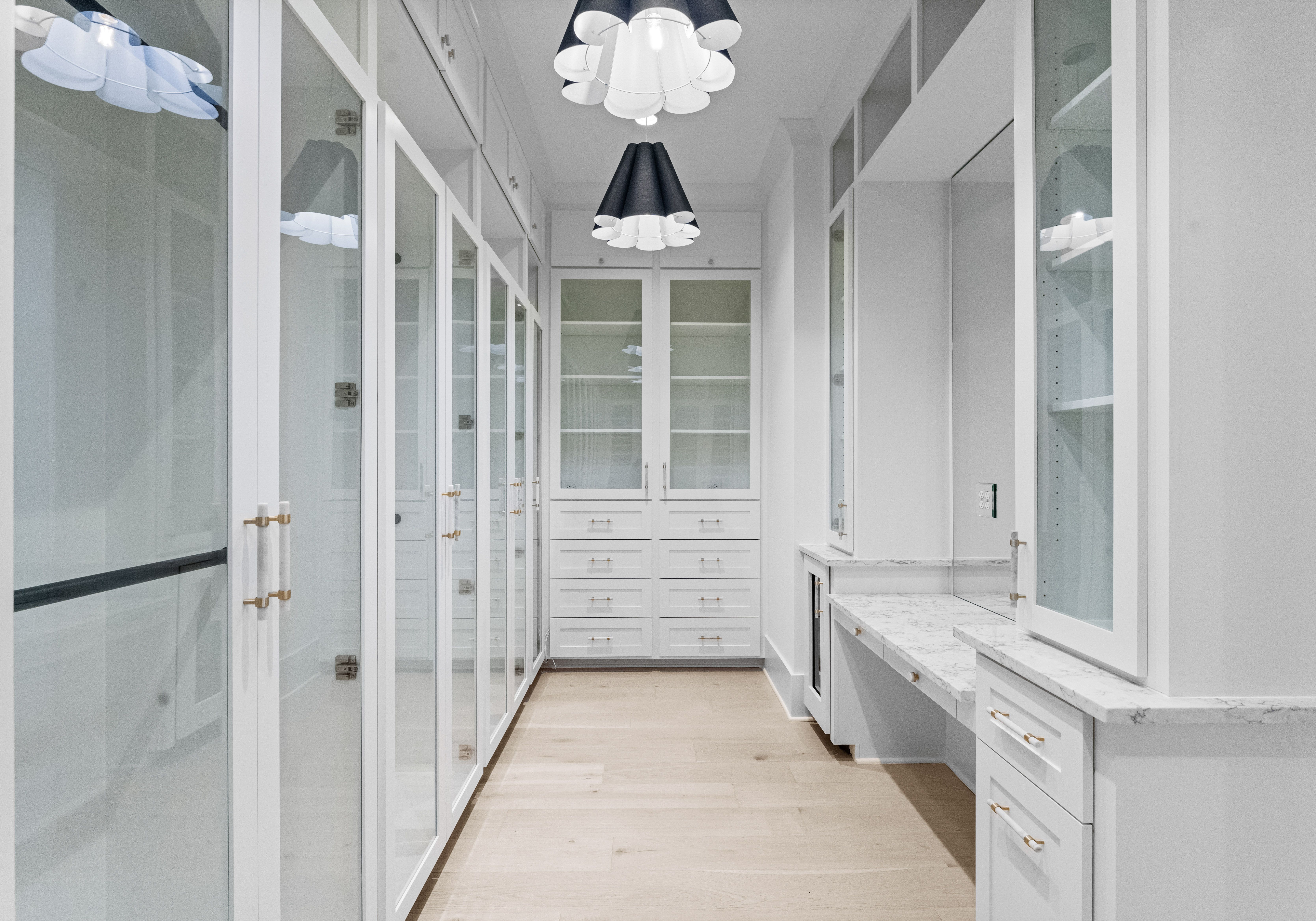 Bright white walk-in closet with glass-front cabinets, light wood floor, marble countertop, and modern black and white pendant lights hanging from ceiling.