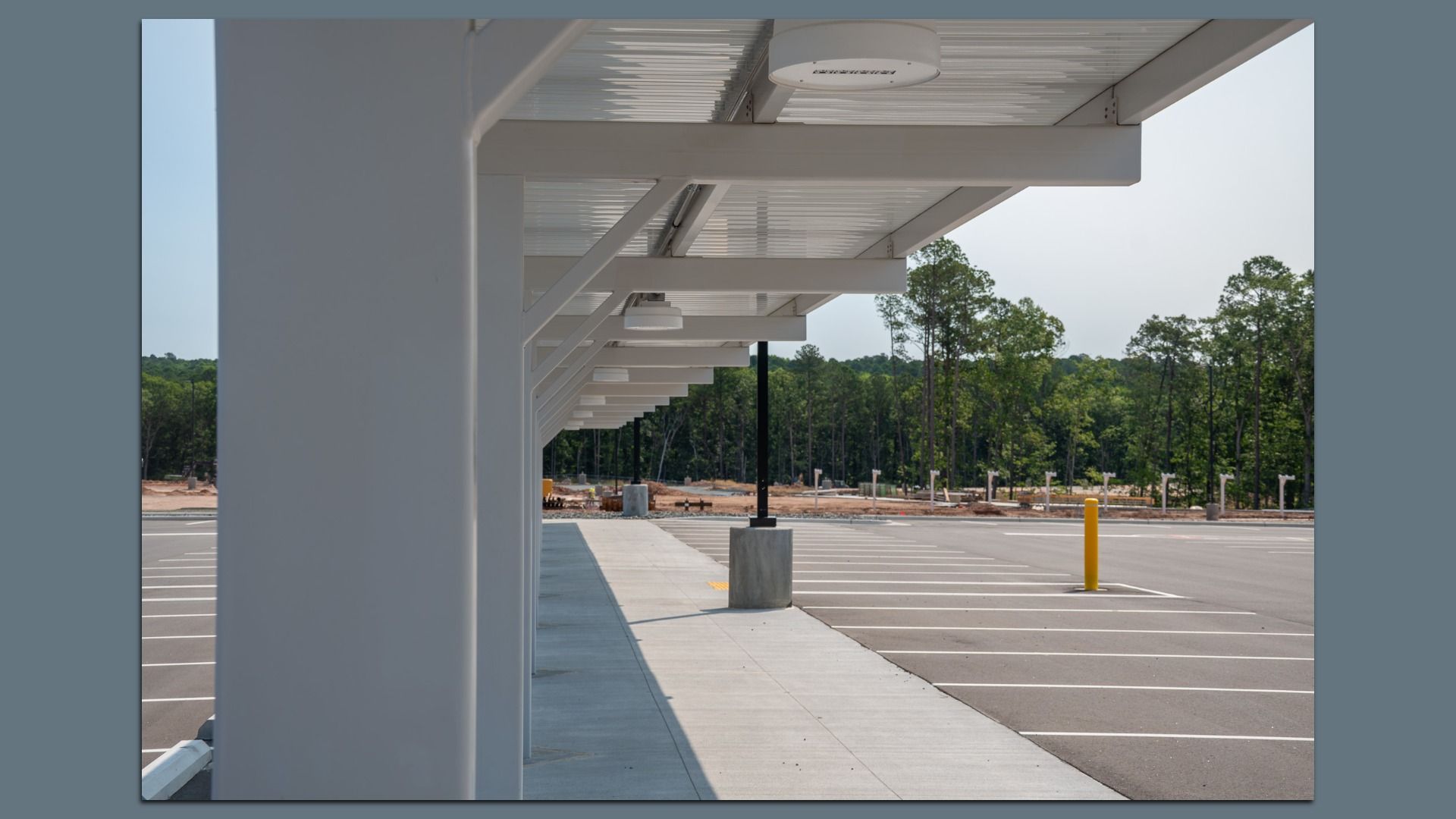 Empty parking lot with white-striped spaces next to a sidewalk covered by a white metal awning with lights, surrounded by green trees under a clear sky.