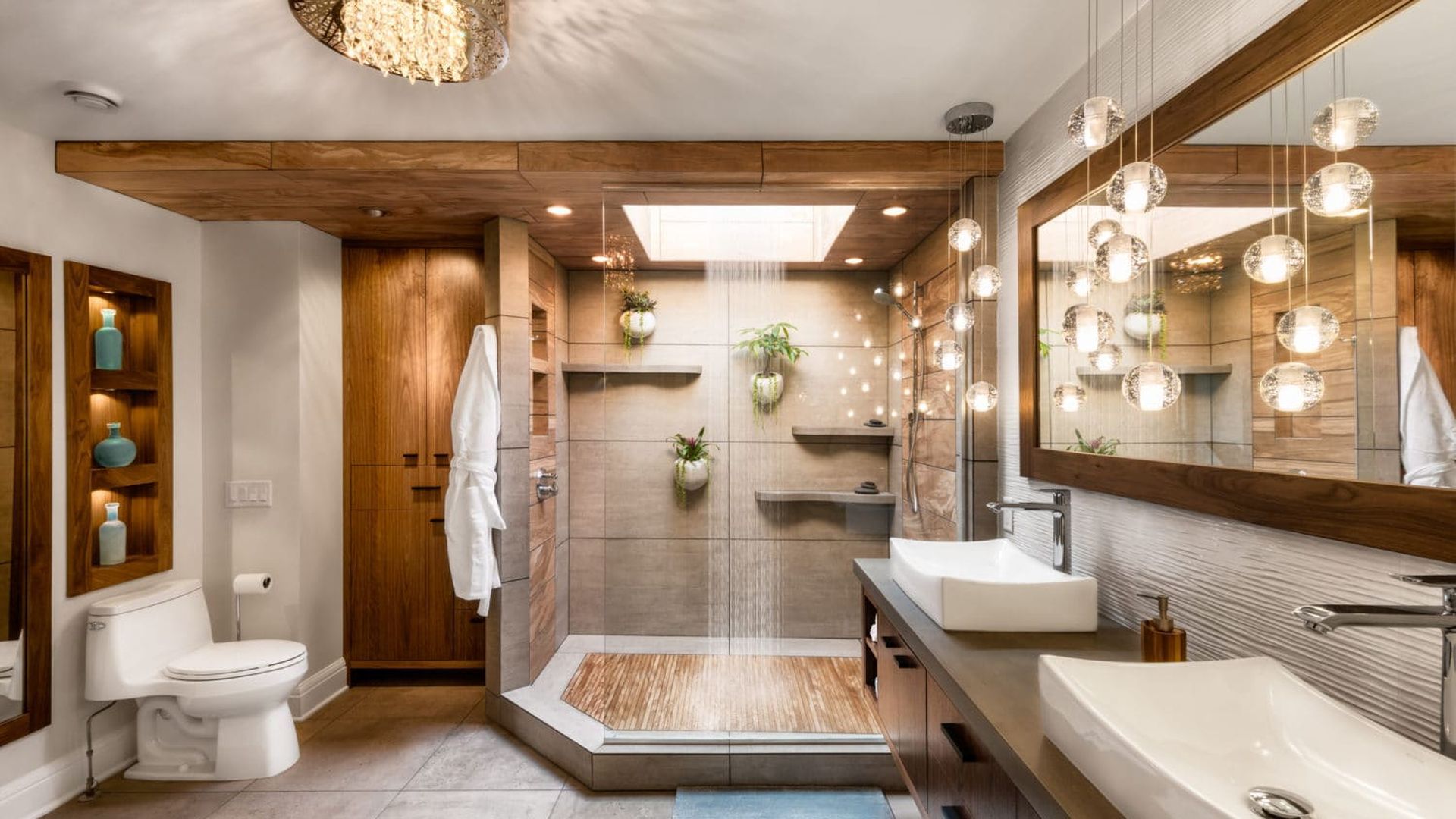 A photo shows a bathroom with lighting in small hanging balls, a chandelier, two sinks, a see-through shower with three plants hanging inside on the wall and a toilet to the left
