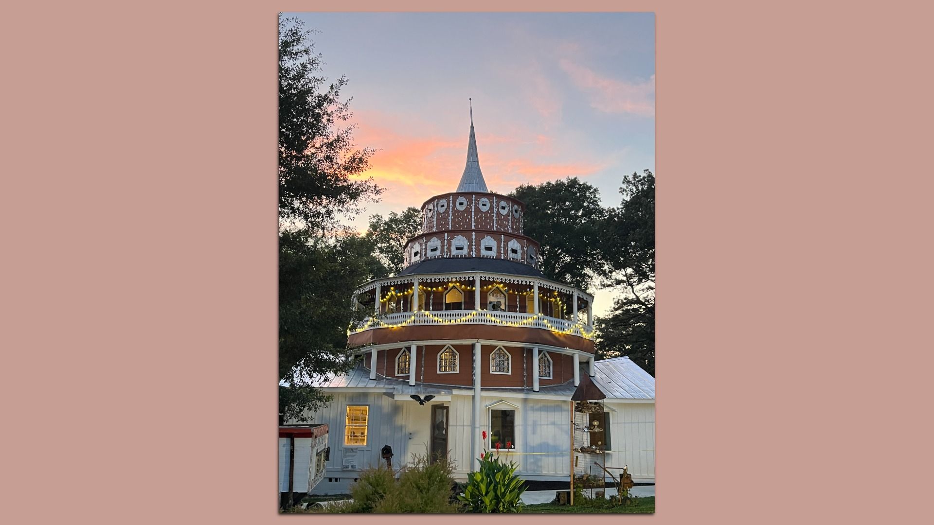 Unique multi-tiered building with round upper levels decorated with white patterns and yellow lights, white lower structure, surrounded by trees, under a pink and blue sunset sky.