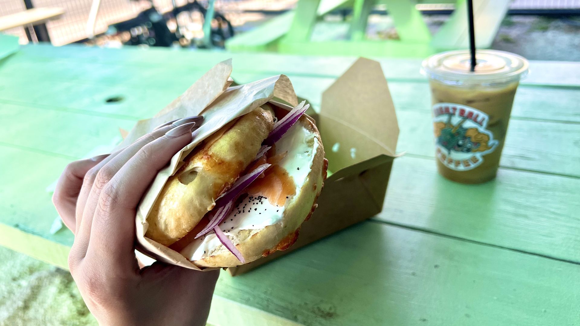 A hand holding a bagel sandwich with cream cheese, smoked salmon, and red onions over a green picnic table with a brown takeout box and iced coffee in a plastic cup with a straw in the background. The bagel is from Bagel Babes and the coffee is from Eight Ball Coffee.